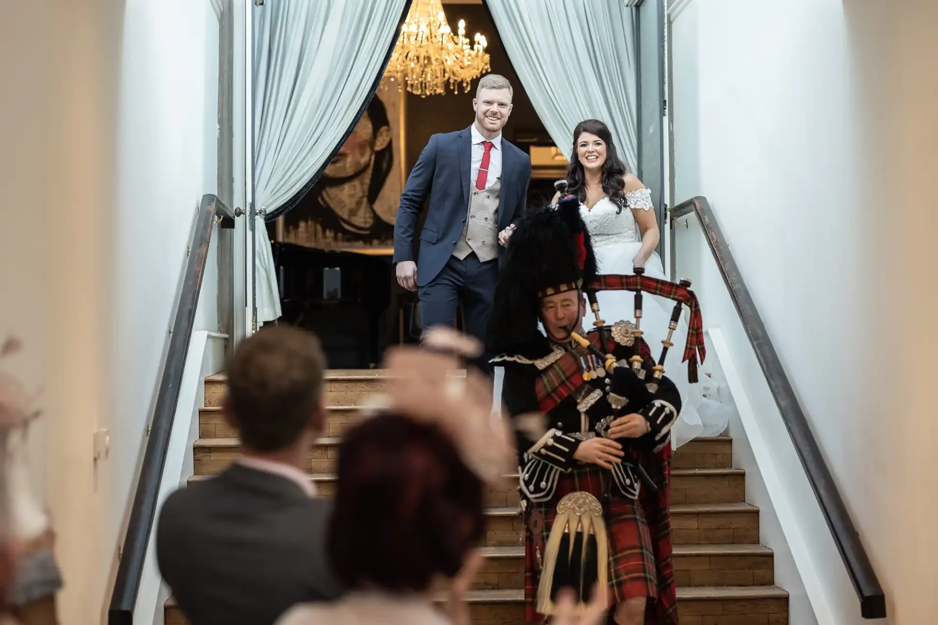 A bride and groom smiling as they descend a staircase with a bagpiper in scottish attire playing at the foreground, with guests clapping.