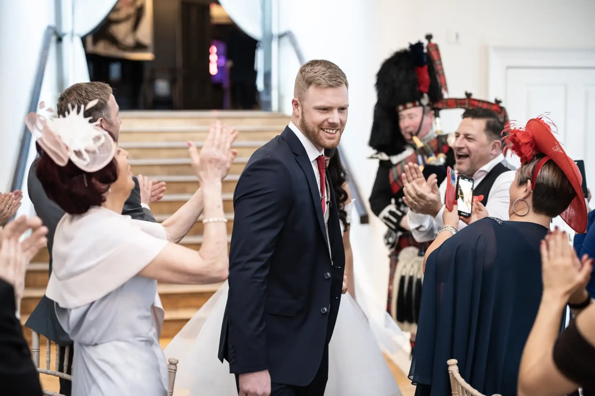 A groom walks down the aisle smiling, greeted by applauding guests in formal attire and two bagpipers in traditional scottish dress.