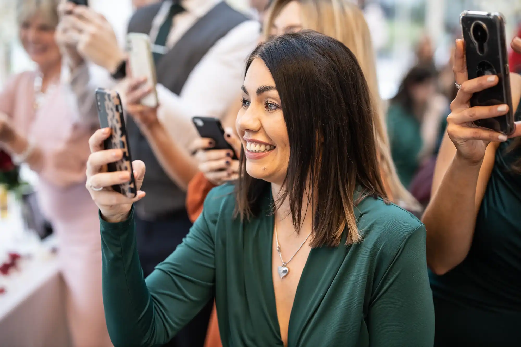 A woman in a green dress smiles while taking a photo with her phone at an event. Other people in the background are also using their phones.