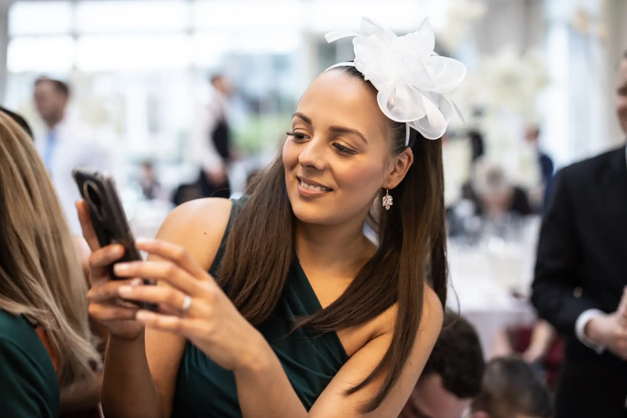 A woman in a green dress with a white bow on her head is holding a phone and smiling in a bright, indoor setting. Other people are visible in the background.