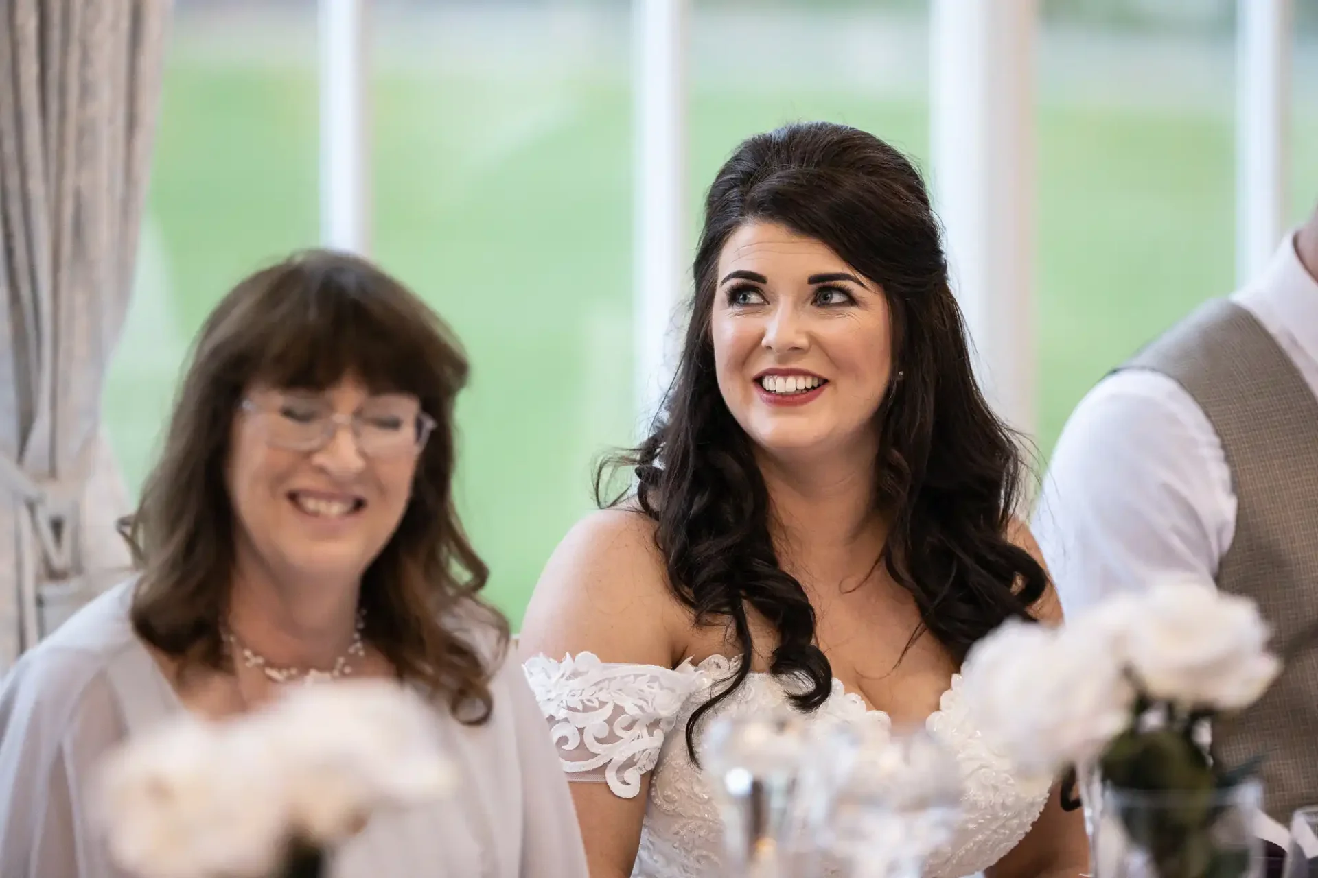 A bride smiling joyfully at a wedding reception, seated next to a woman in glasses. they are surrounded by elegant floral decorations.