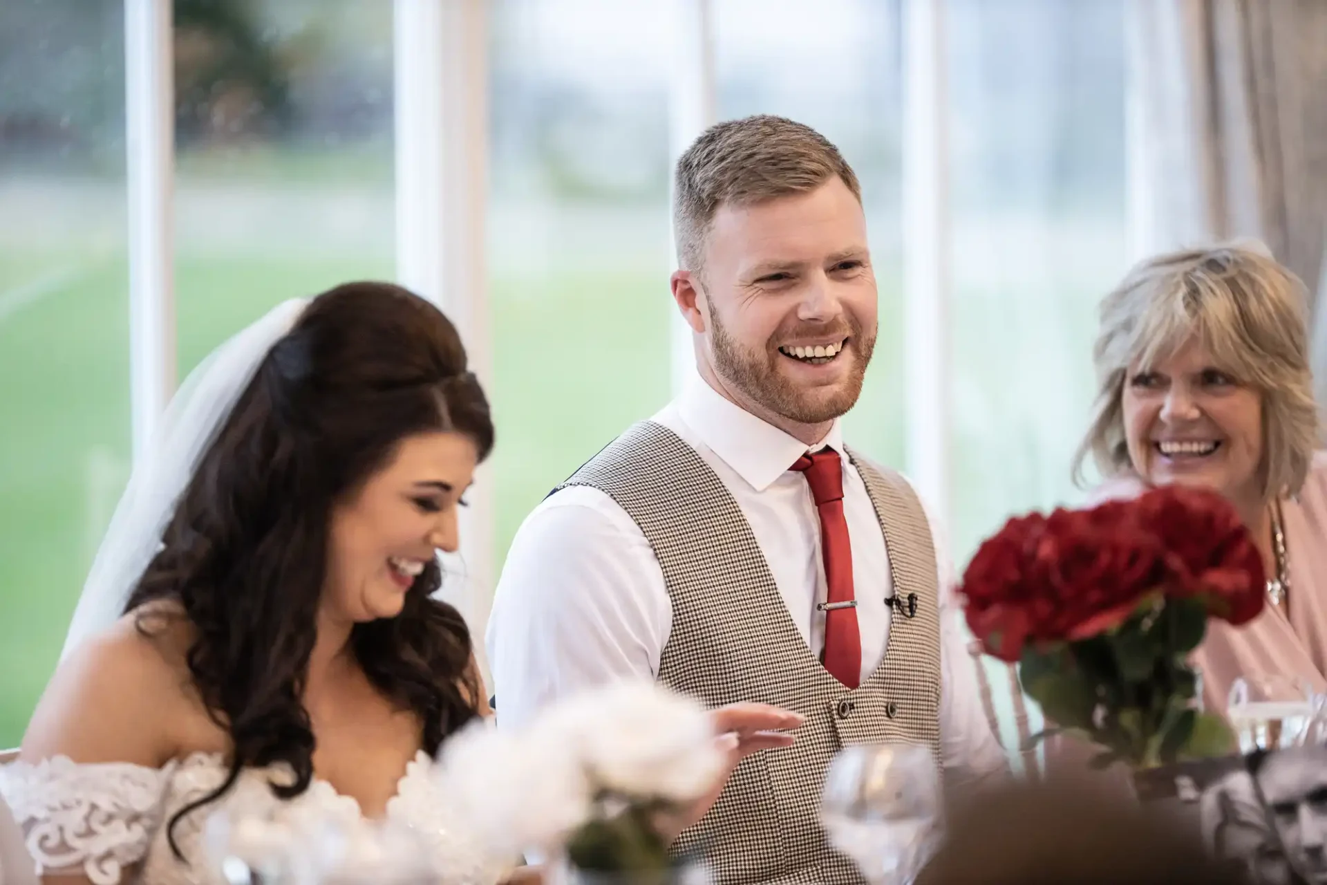 A groom in a tweed vest laughs jovially at a wedding table with a bride in a lace dress and an older woman smiling nearby, with roses and elegant decor around.