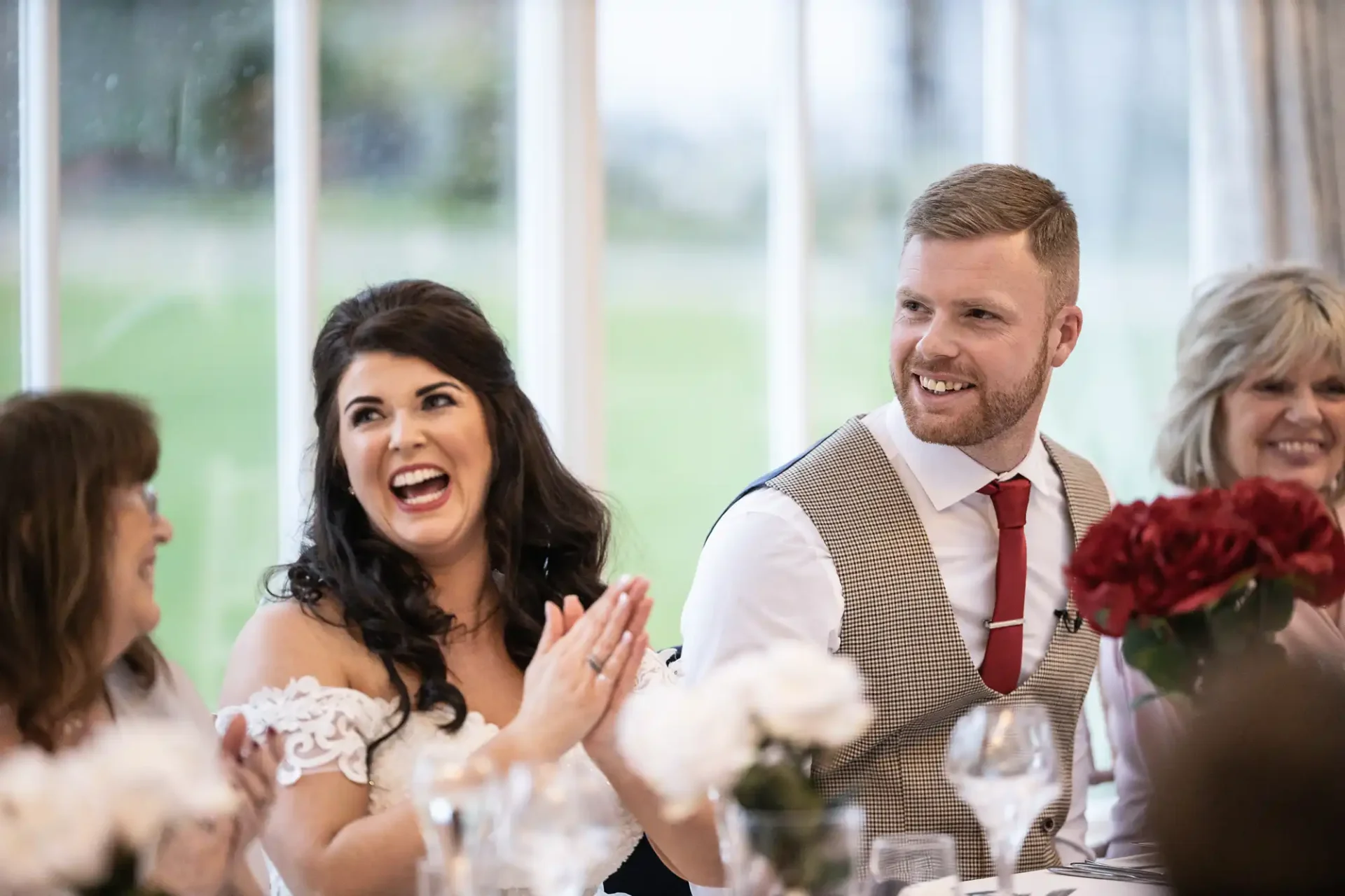 A joyful bride and groom sit at a wedding reception, laughing together in a brightly lit room with large windows overlooking a green landscape.