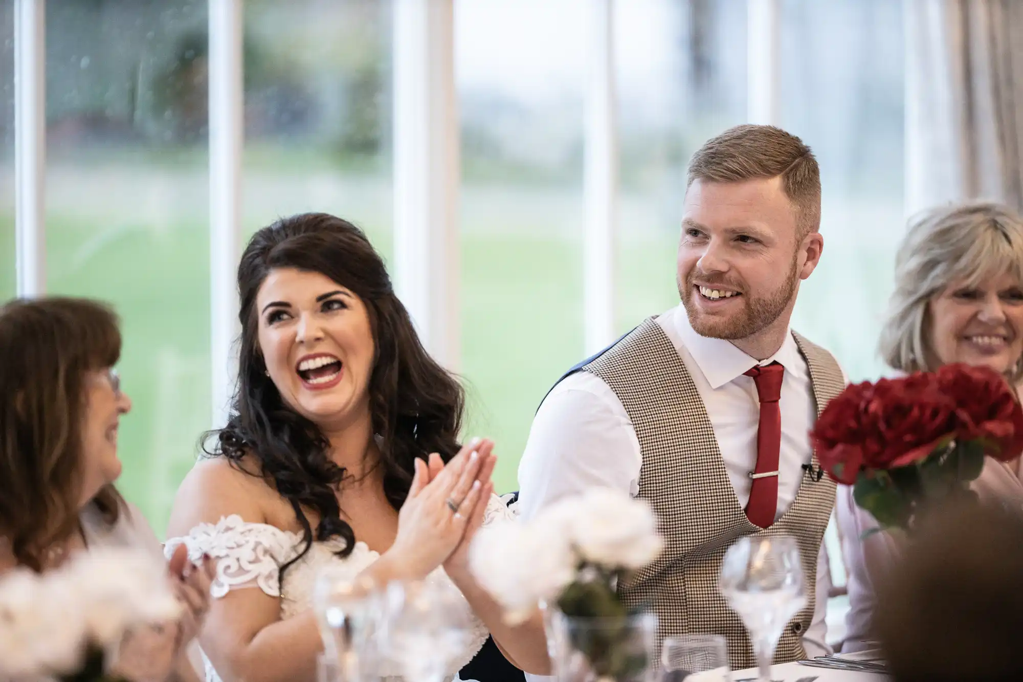 A bride and groom smile and laugh with guests at a table decorated with white flowers and red roses.