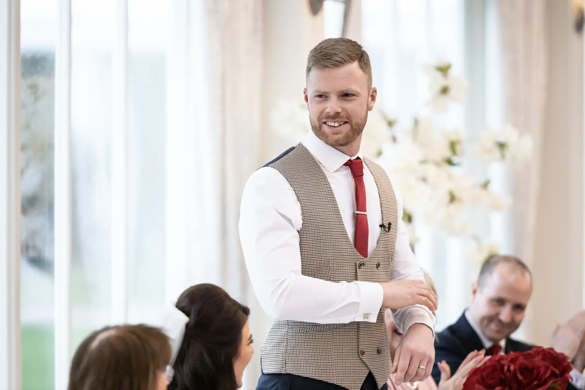 A man wearing a tweed vest and red tie smiles while standing at a formal event, with seated guests blurred in the background.