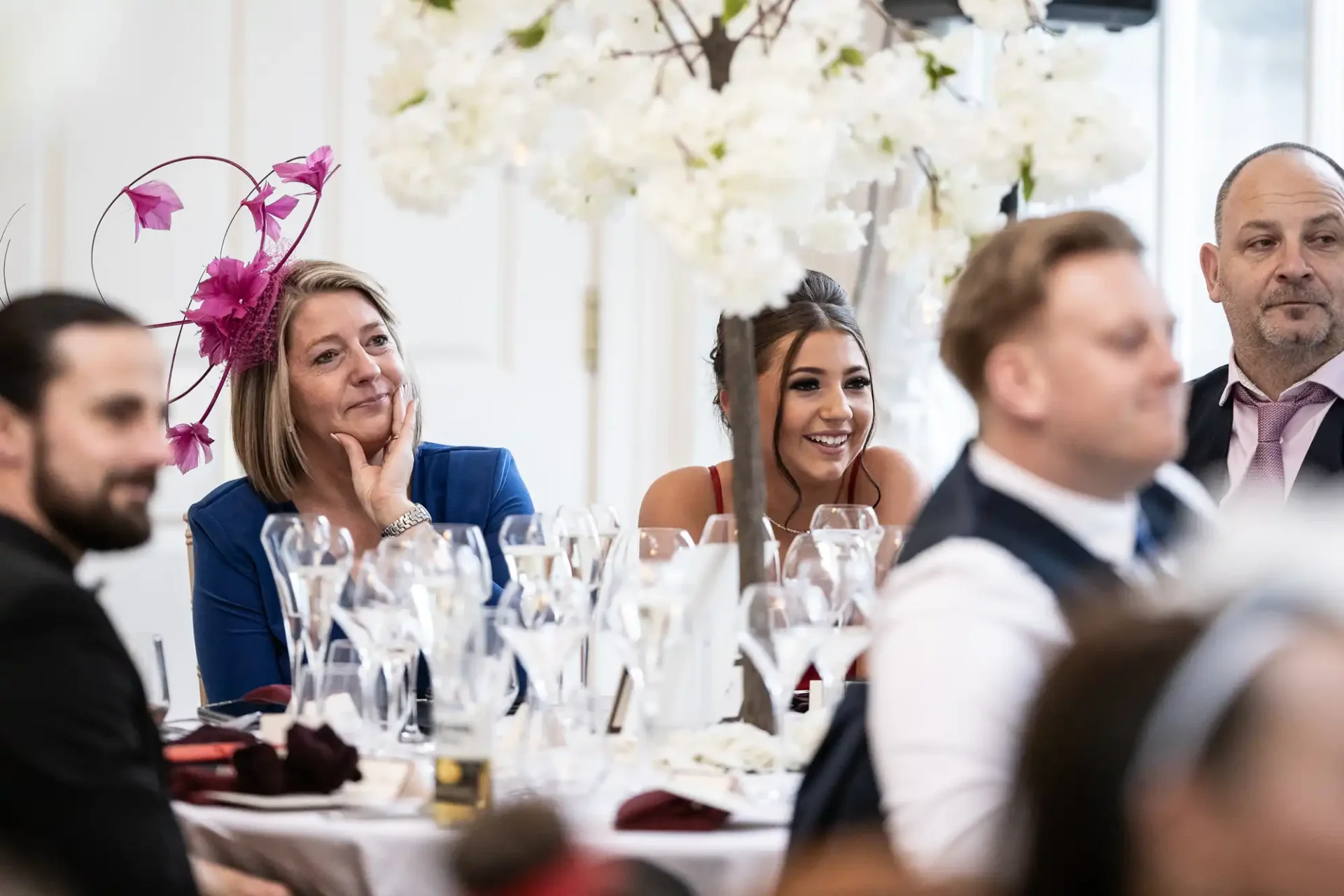 Guests at a wedding reception are seated at a table adorned with tall white floral centerpieces, with one woman wearing a striking pink fascinator.