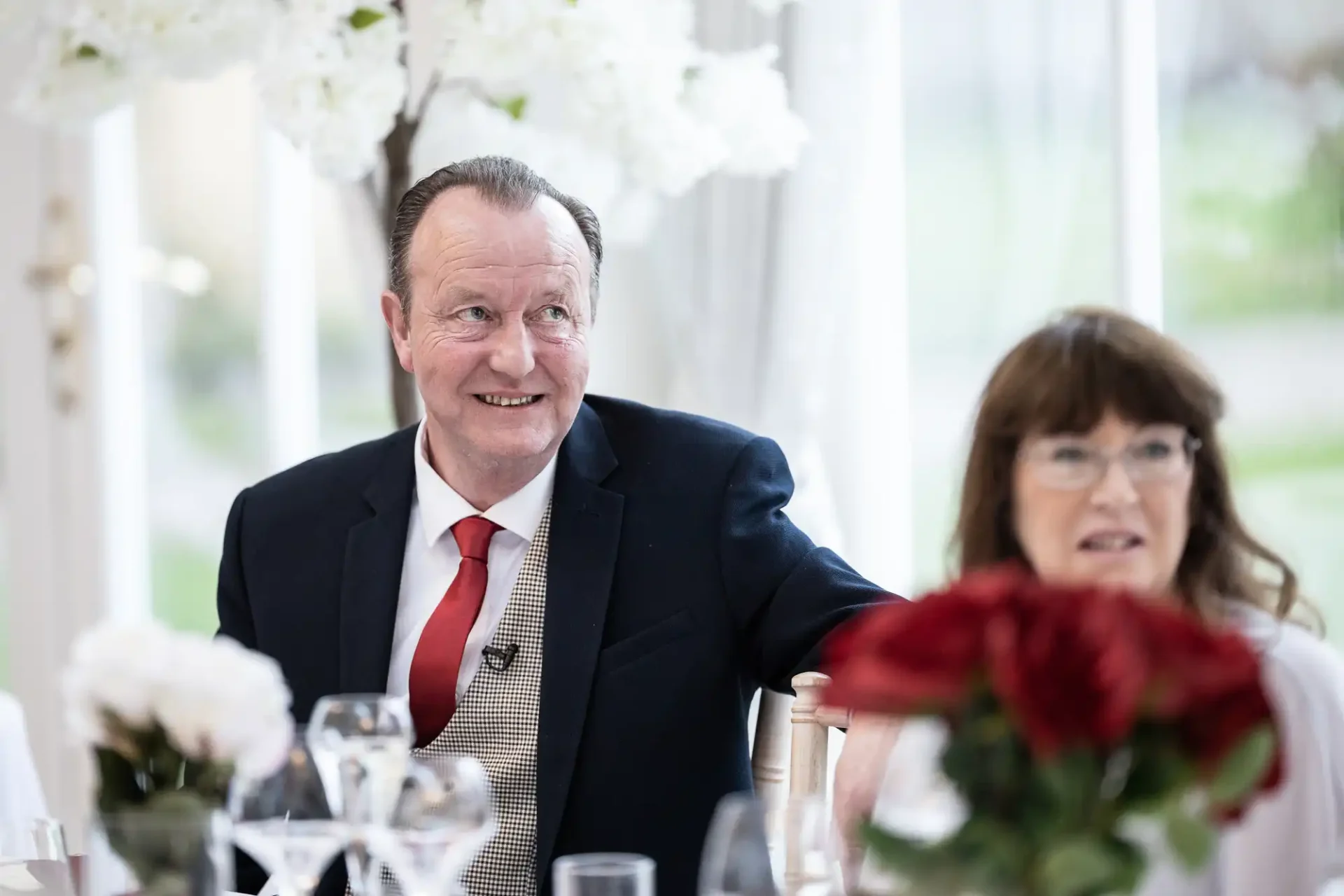 Older man in a suit and tie smiling at a social event, sitting beside a woman with a floral centerpiece in foreground.