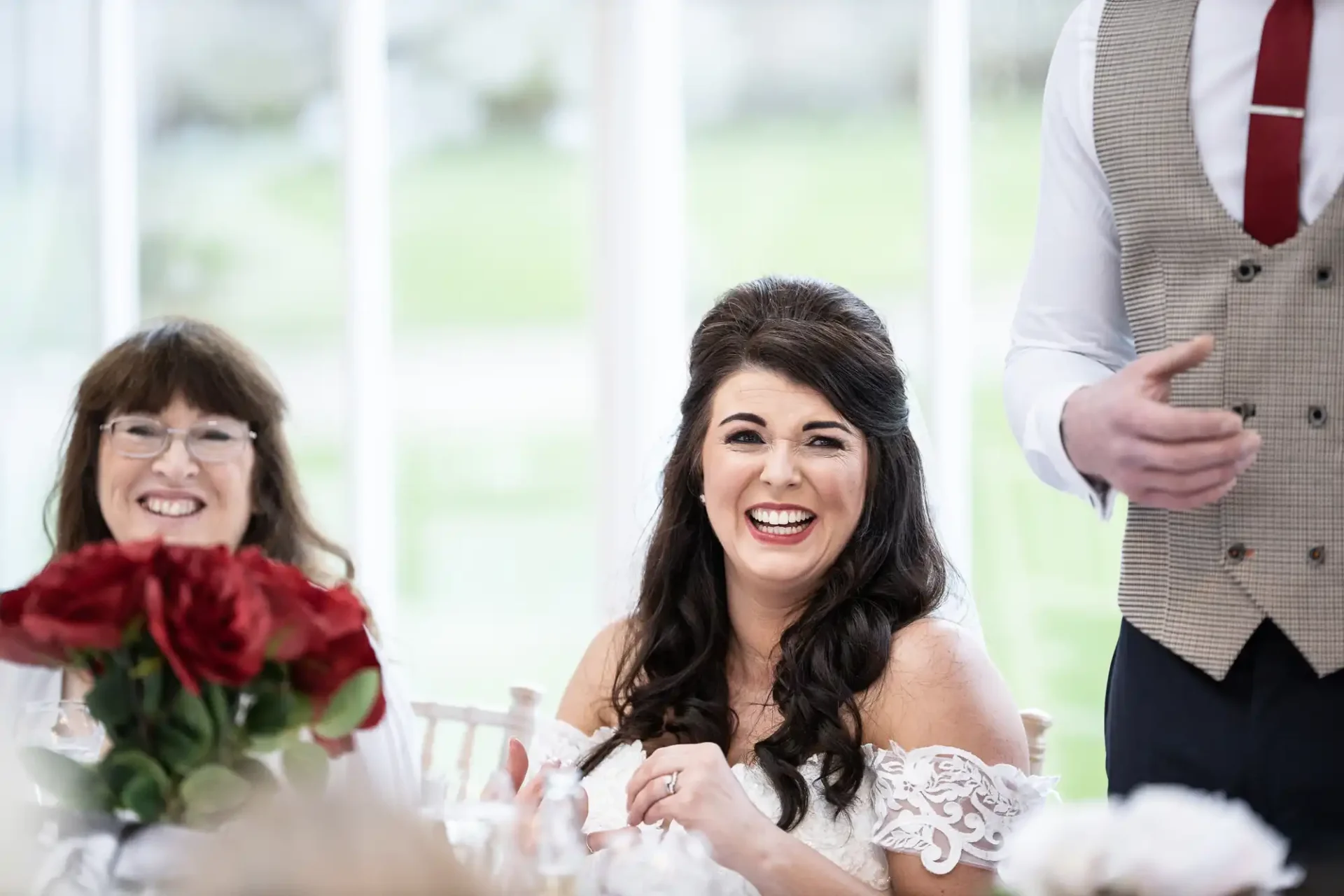 A bride laughing joyfully at a wedding reception table, with a woman smiling beside her and a man gesturing, in a room with bright windows.