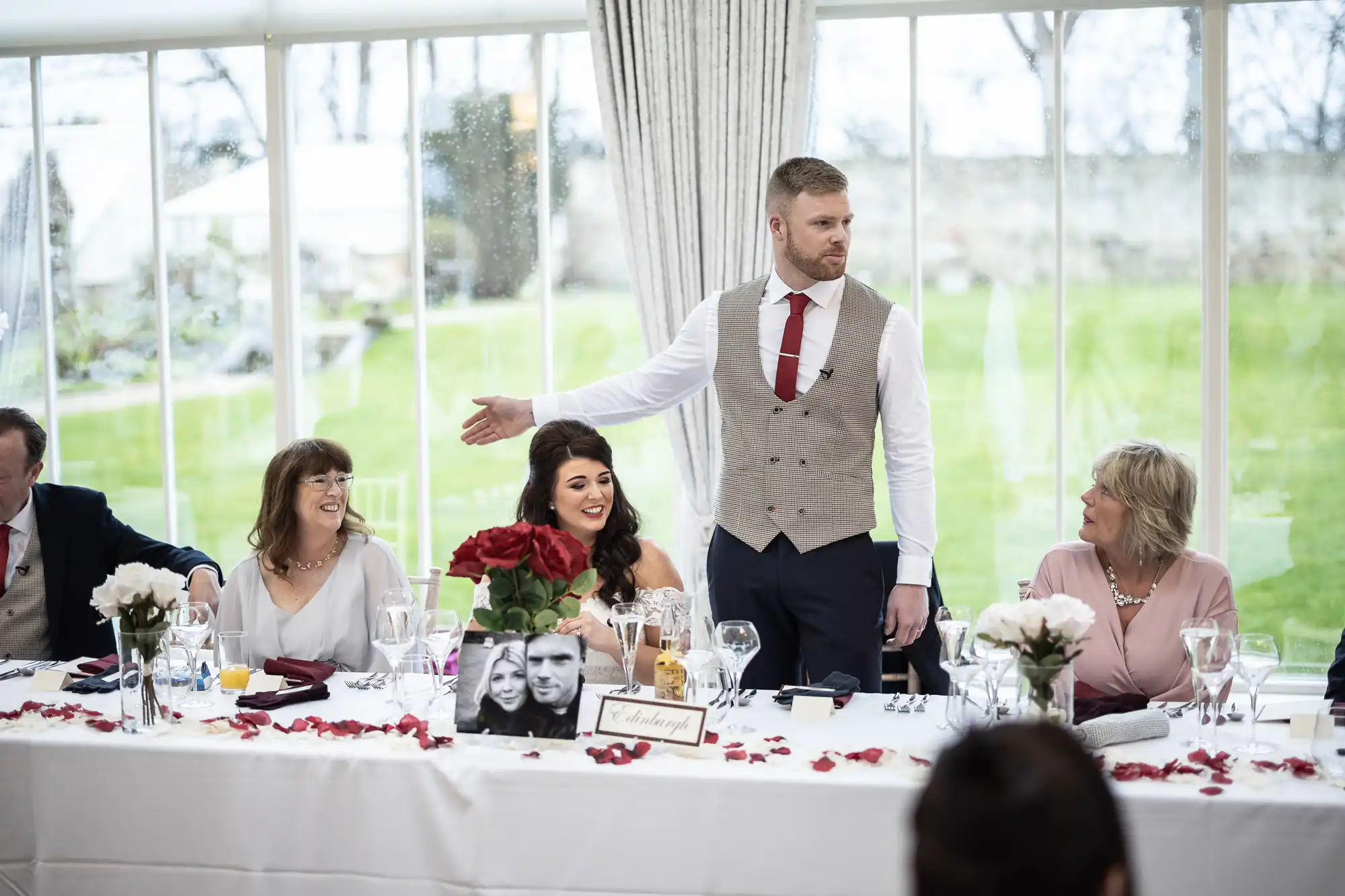 A man in a vest and tie gives a speech at a table set for a formal event. Three women and another man are seated, listening and smiling. The table is decorated with flowers and framed photos.