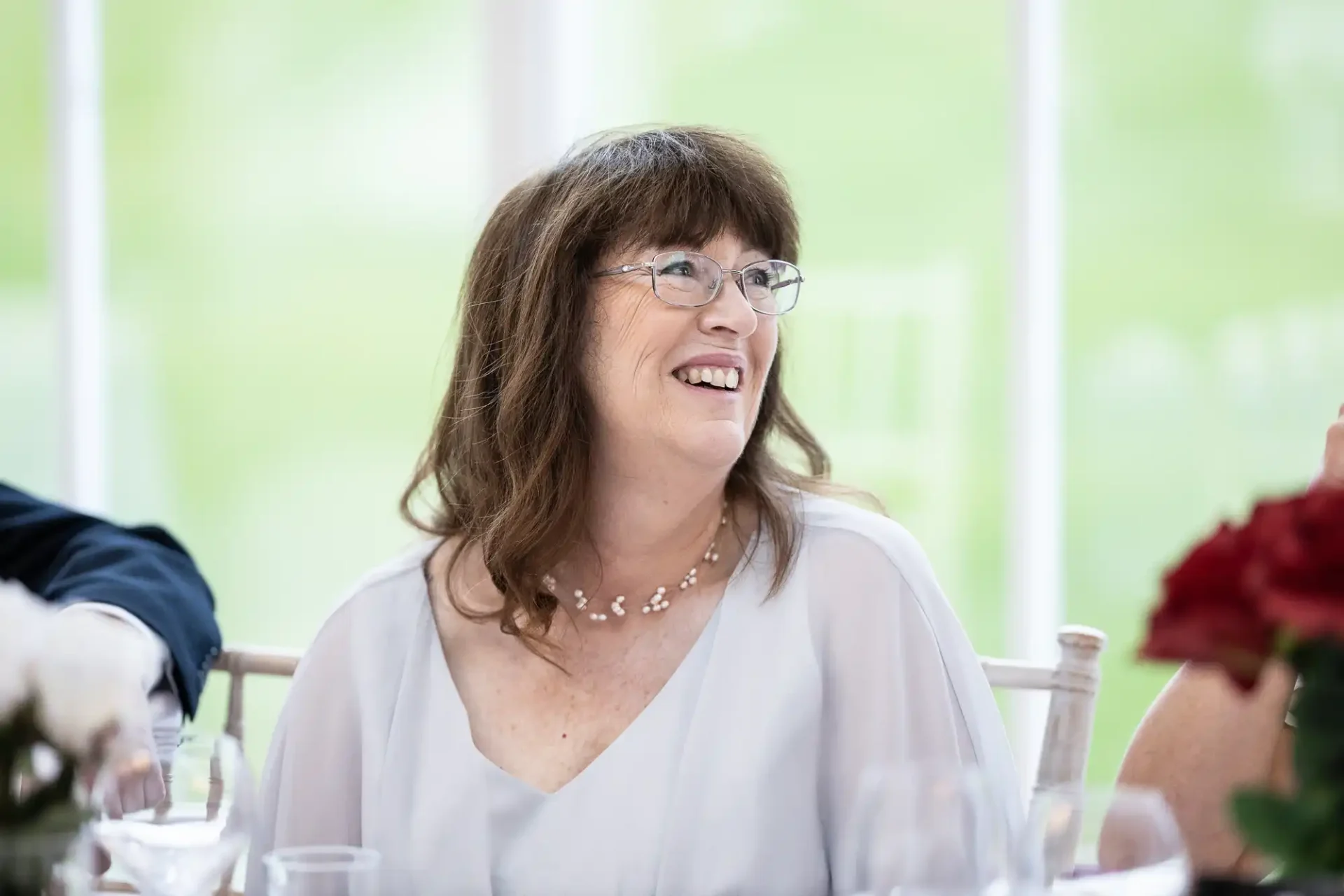 A middle-aged woman with brown hair and glasses, wearing a white blouse, smiles while seated at a table during an event.