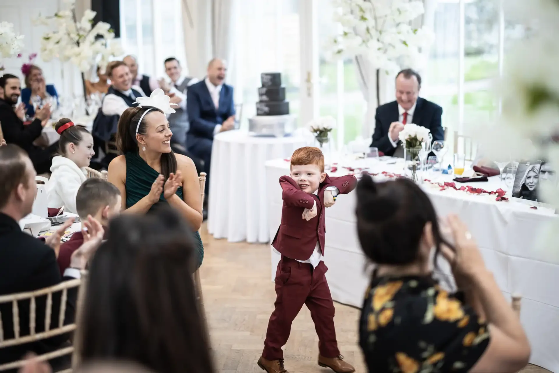 A young boy in a red suit playfully dances at a wedding reception, surrounded by applauding guests.