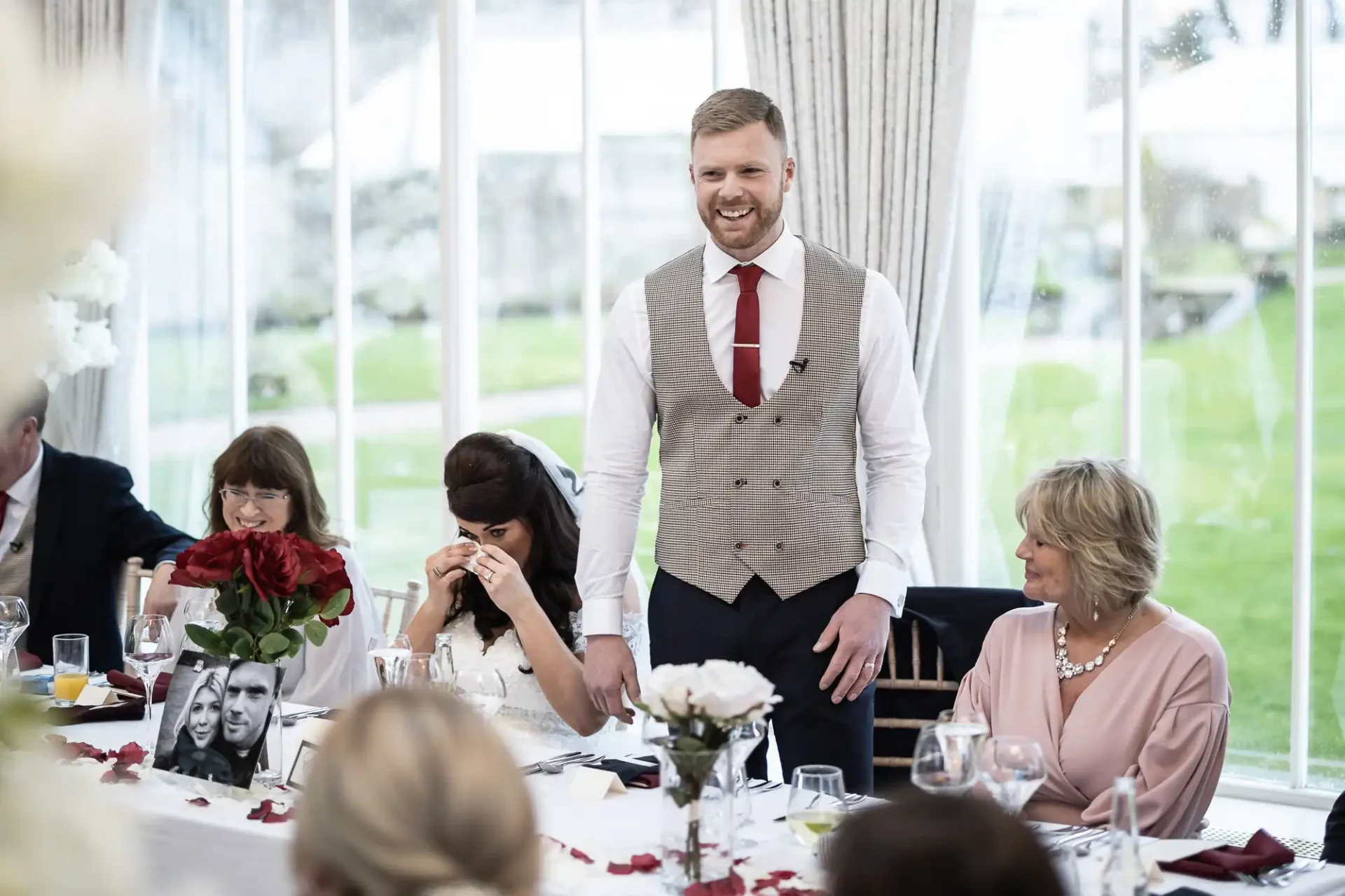 Man in a vest and tie standing and speaking at a formal dining event, with seated guests listening, some smiling.