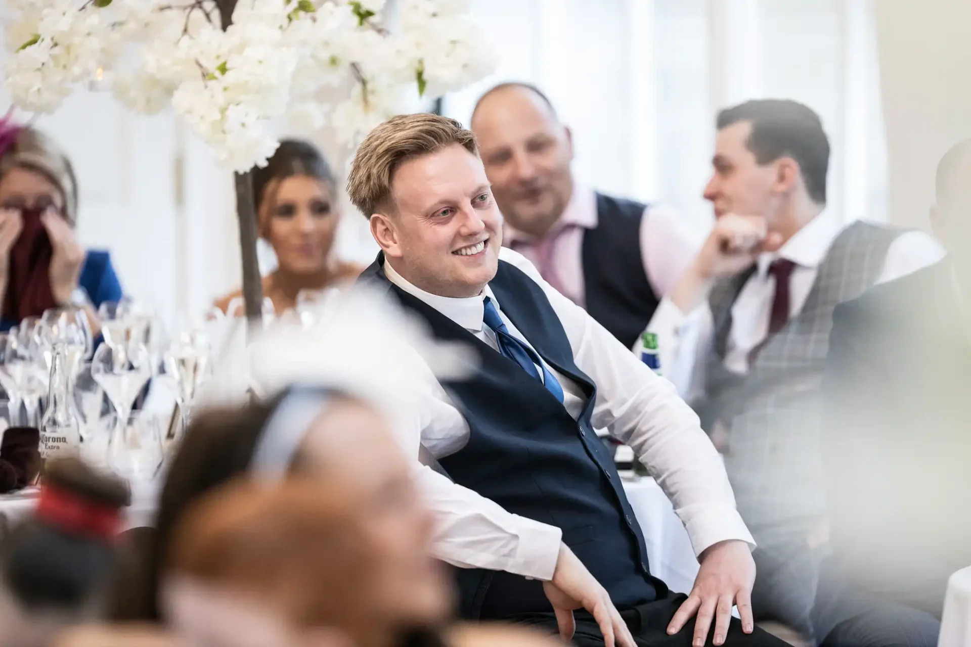 A smiling man in a vest at a wedding reception, surrounded by guests, with focus on his joyful expression.