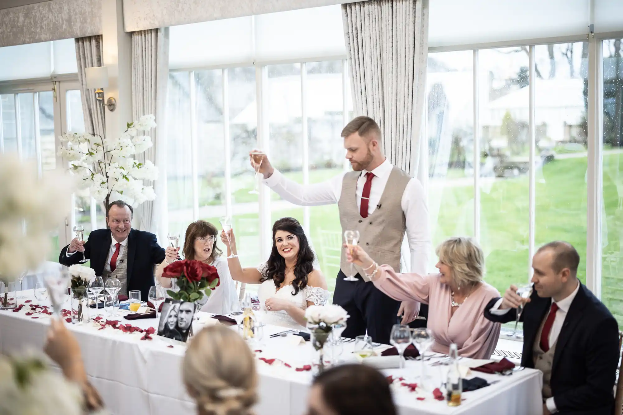 A wedding reception scene with six people seated at a long table. The bride and groom, along with the guests, are raising their glasses in a toast.