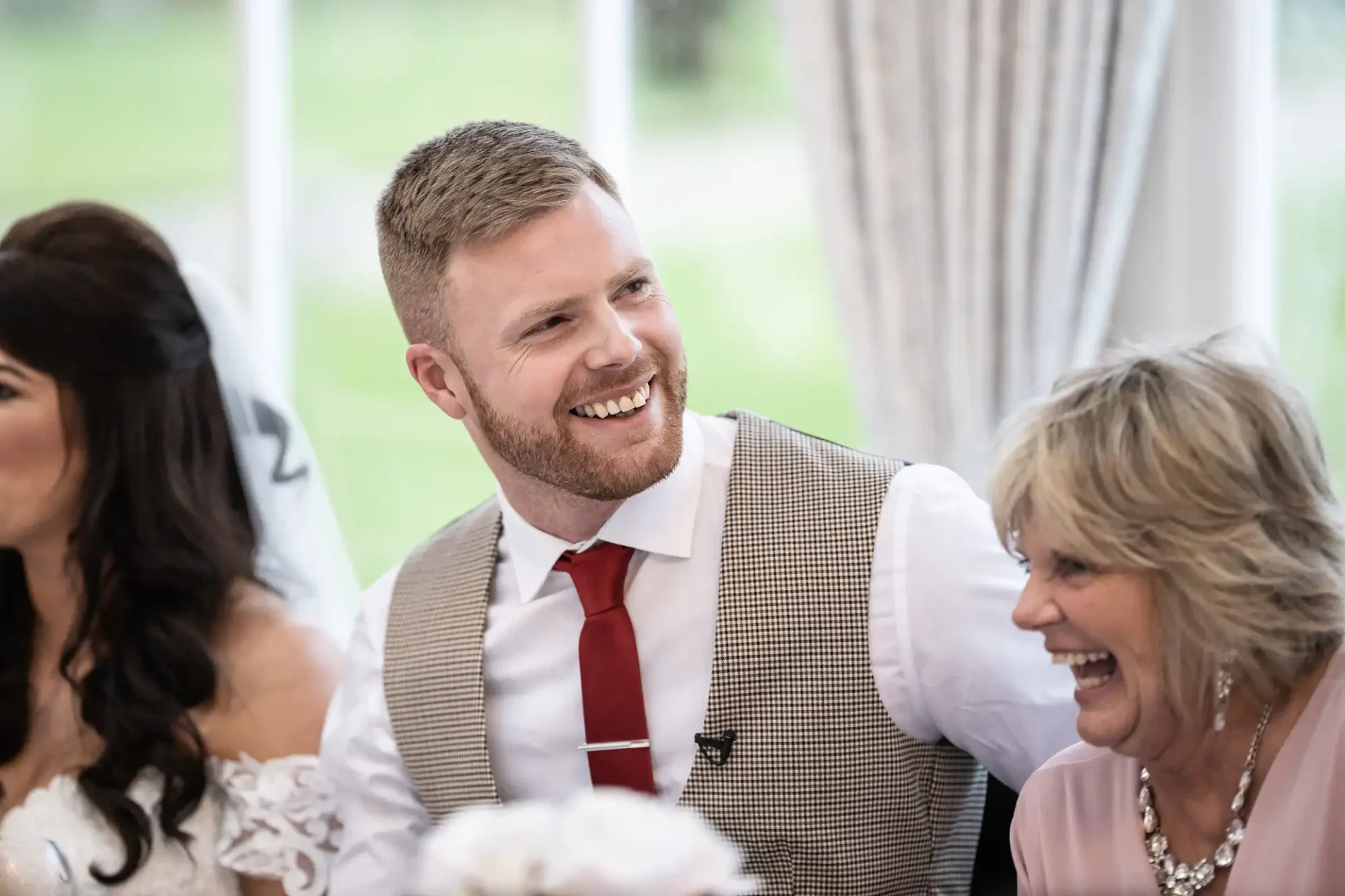 A man in a tweed vest and red tie smiles joyfully at a wedding, sitting beside a bride and an older woman who is laughing.