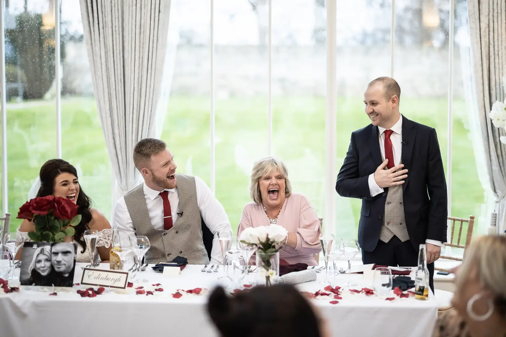 A man in a red tie standing and laughing at a wedding table with three seated guests who are also laughing joyously, surrounded by elegant decor and large windows.