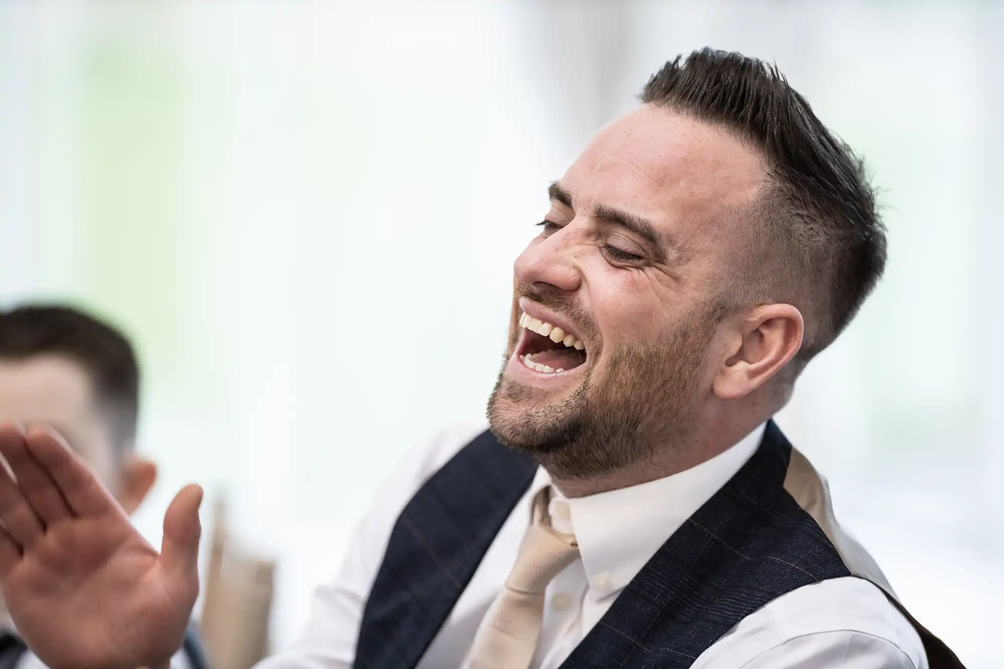 A man wearing a suit with a vest and tie is laughing heartily, with his mouth open and eyes closed.