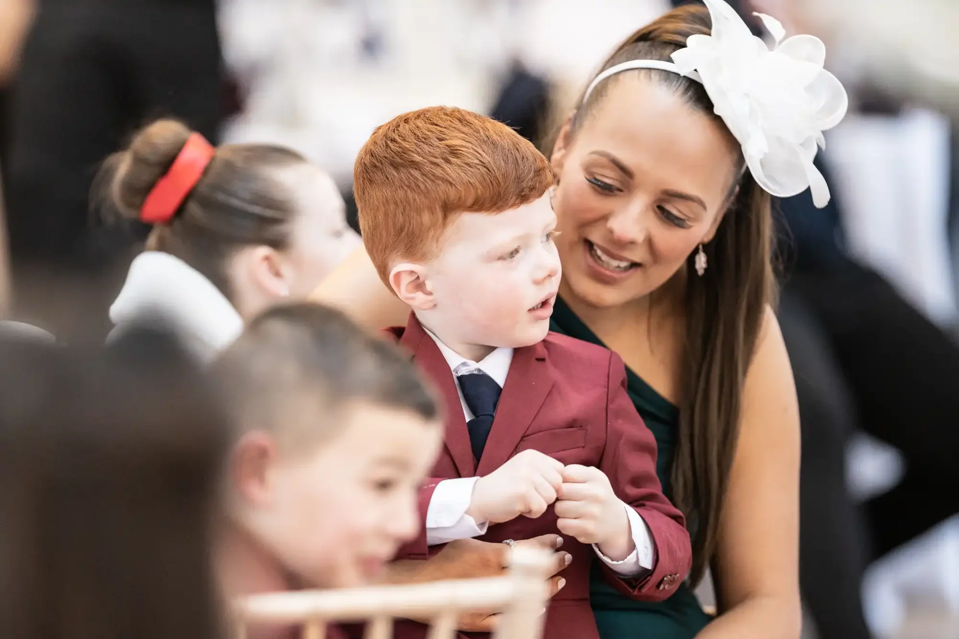 A young boy with red hair, dressed in a burgundy suit, converses with a smiling woman wearing a white fascinator at a formal event.