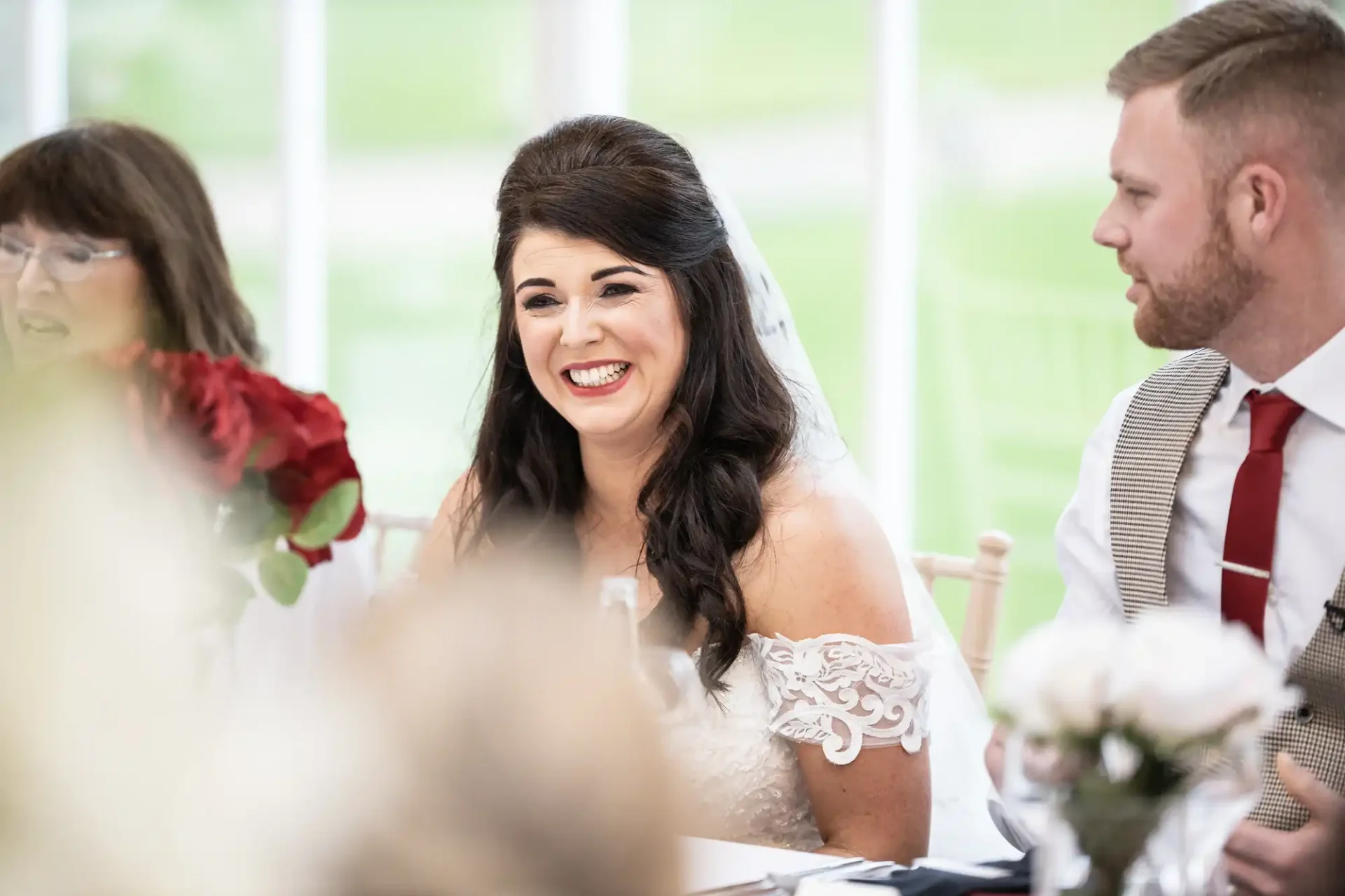 A bride in a white off-shoulder wedding dress laughs joyfully at a table during a wedding reception, with guests nearby.