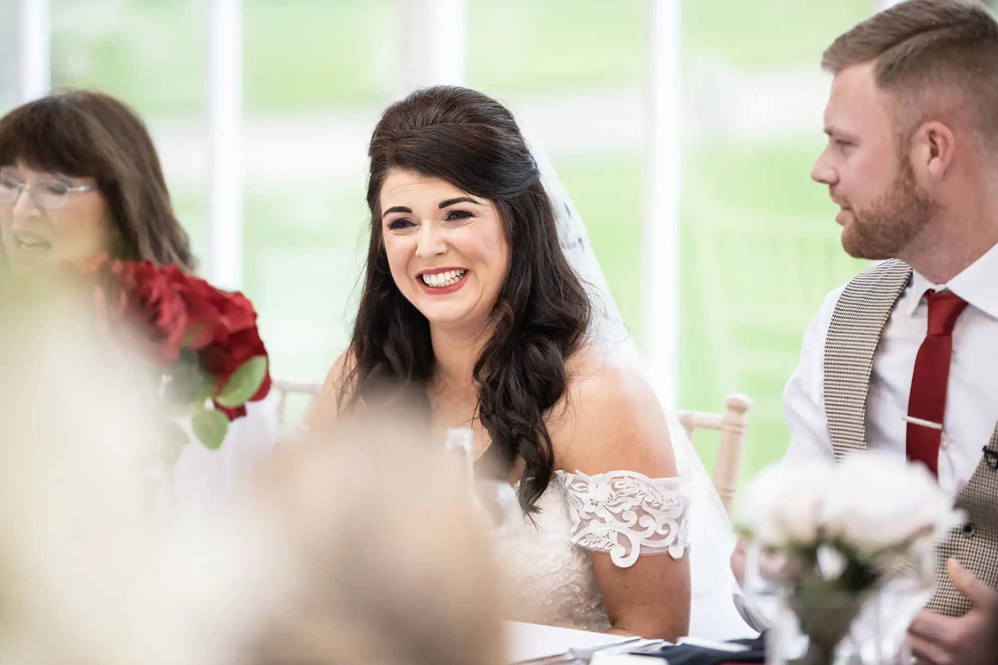 Bride in a white dress smiles while seated at a table between a man in a vest and tie and another woman, with bouquets of flowers in the foreground.
