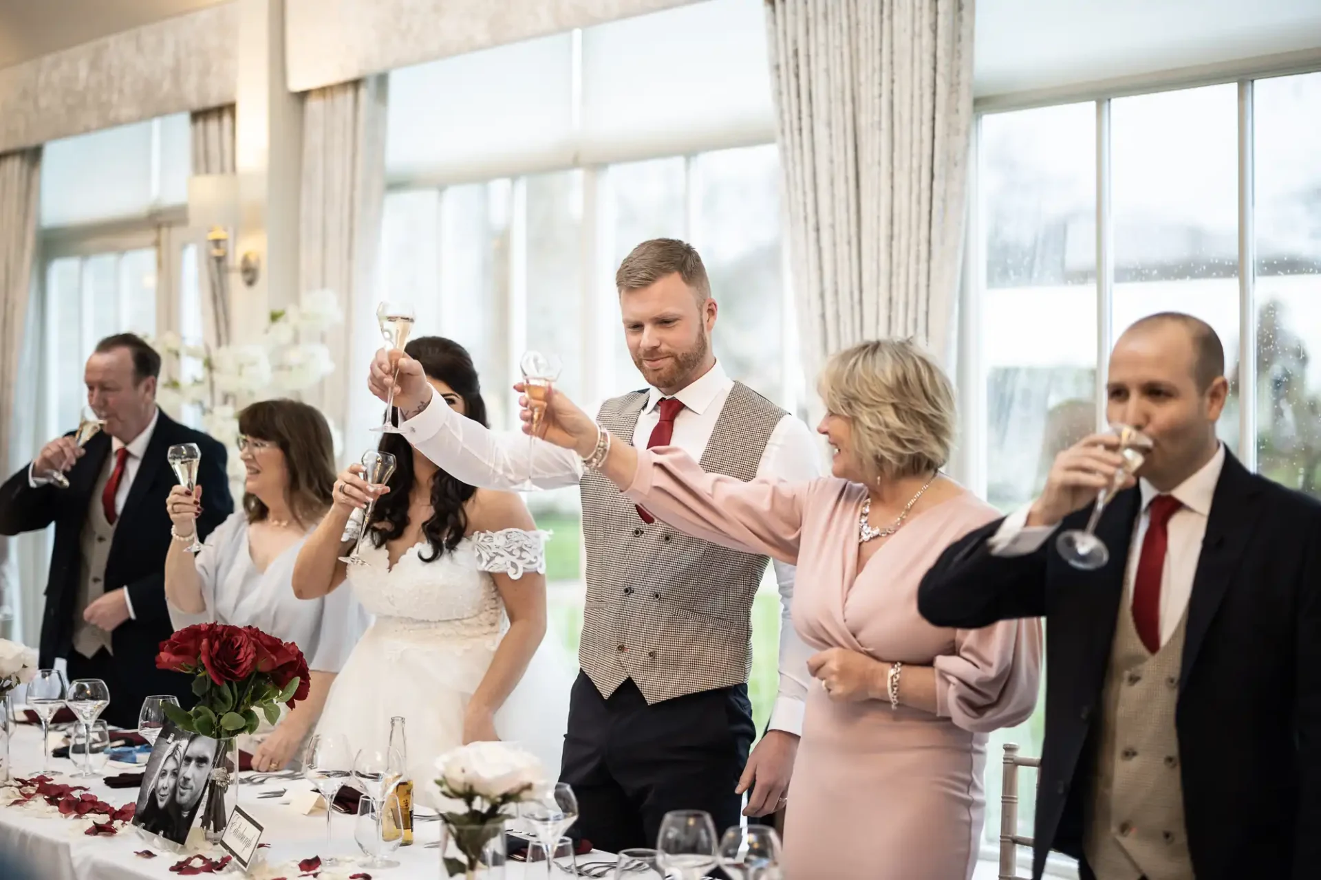 A wedding toast inside a banquet hall, where a groom and several guests raise glasses with a window view background.