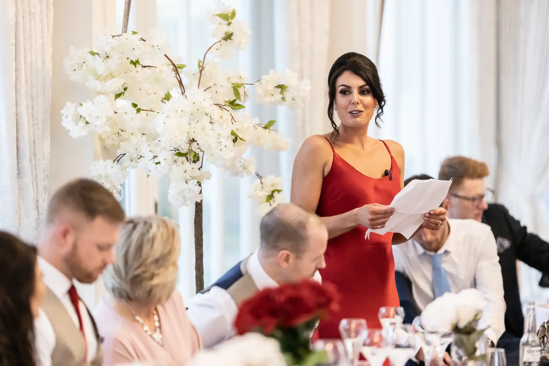 Woman in red dress giving a speech at a wedding reception, holding a paper, with guests seated at tables listening.