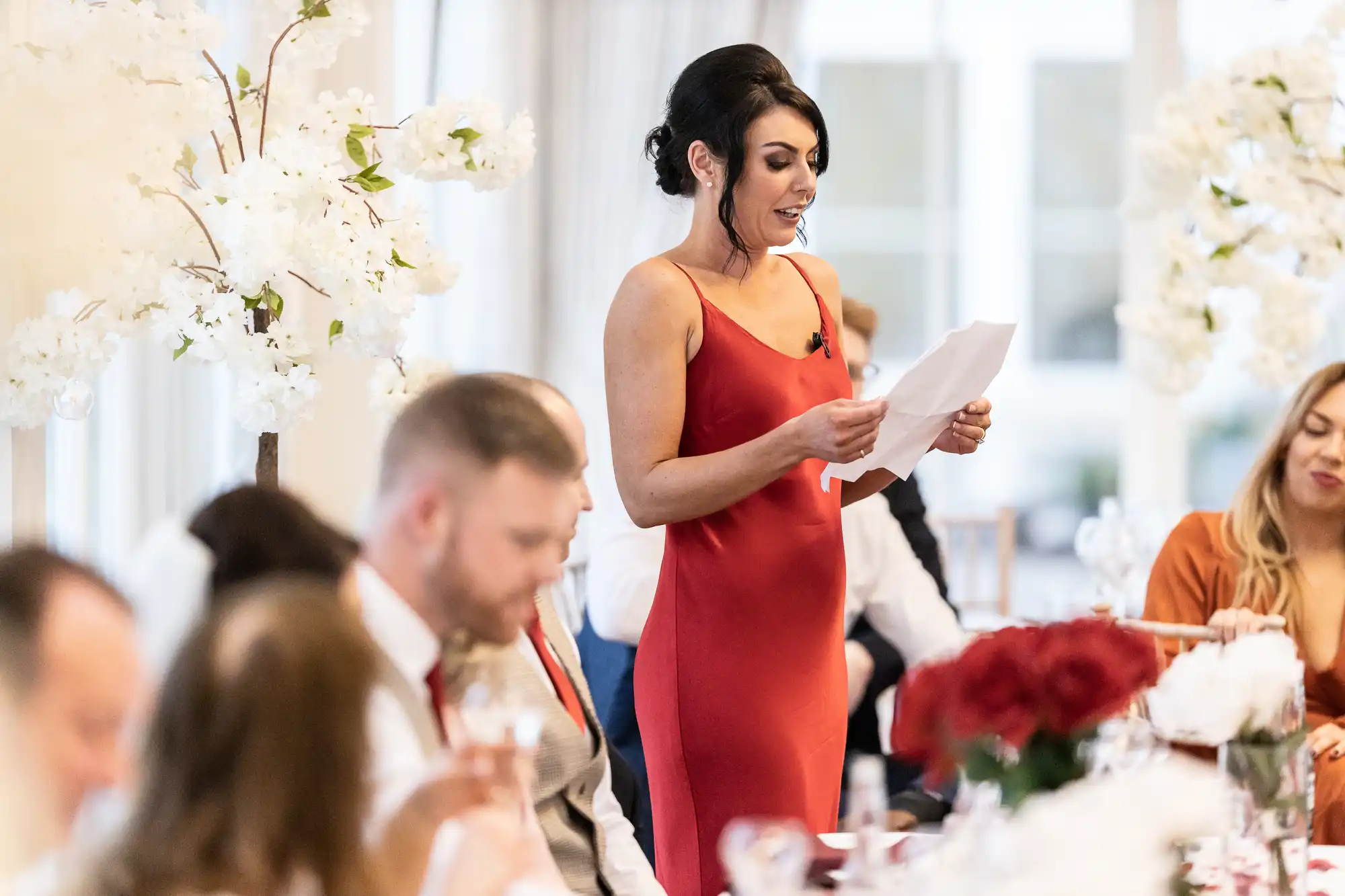 A woman in a red dress is giving a speech while standing among seated guests at a formal event decorated with white flowers.