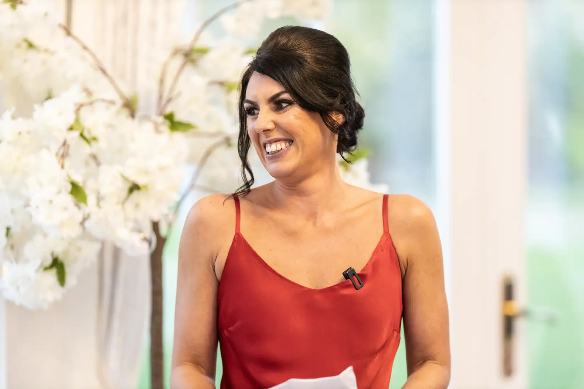 A woman in a red top smiles while looking to the side, standing indoors in a room decorated with white flowers.