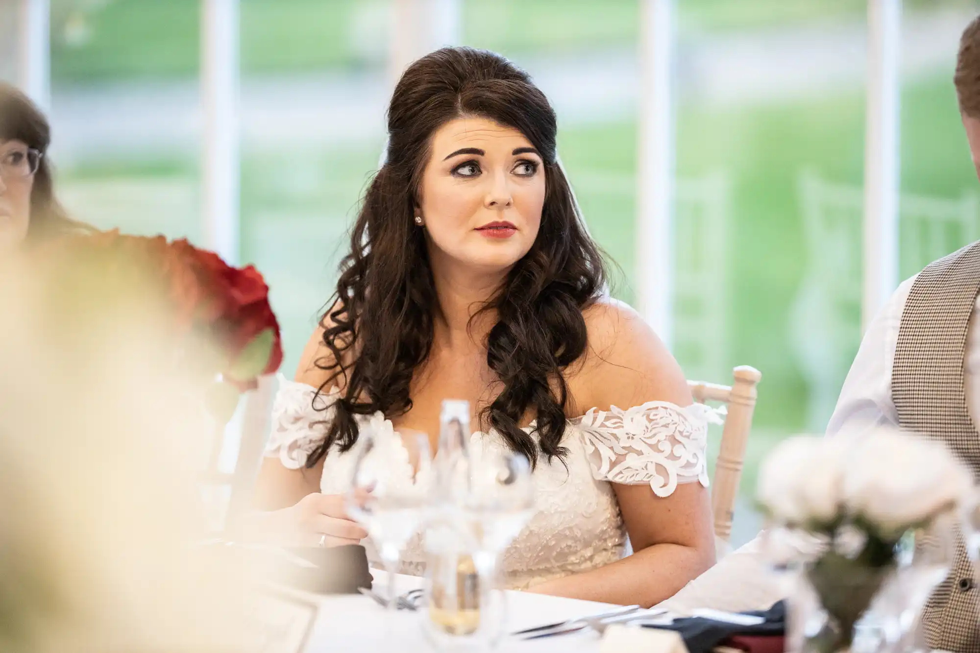A woman in a white wedding dress sits at a table, looking to her left with a pensive expression. Glasses and flowers are on the table in front of her.