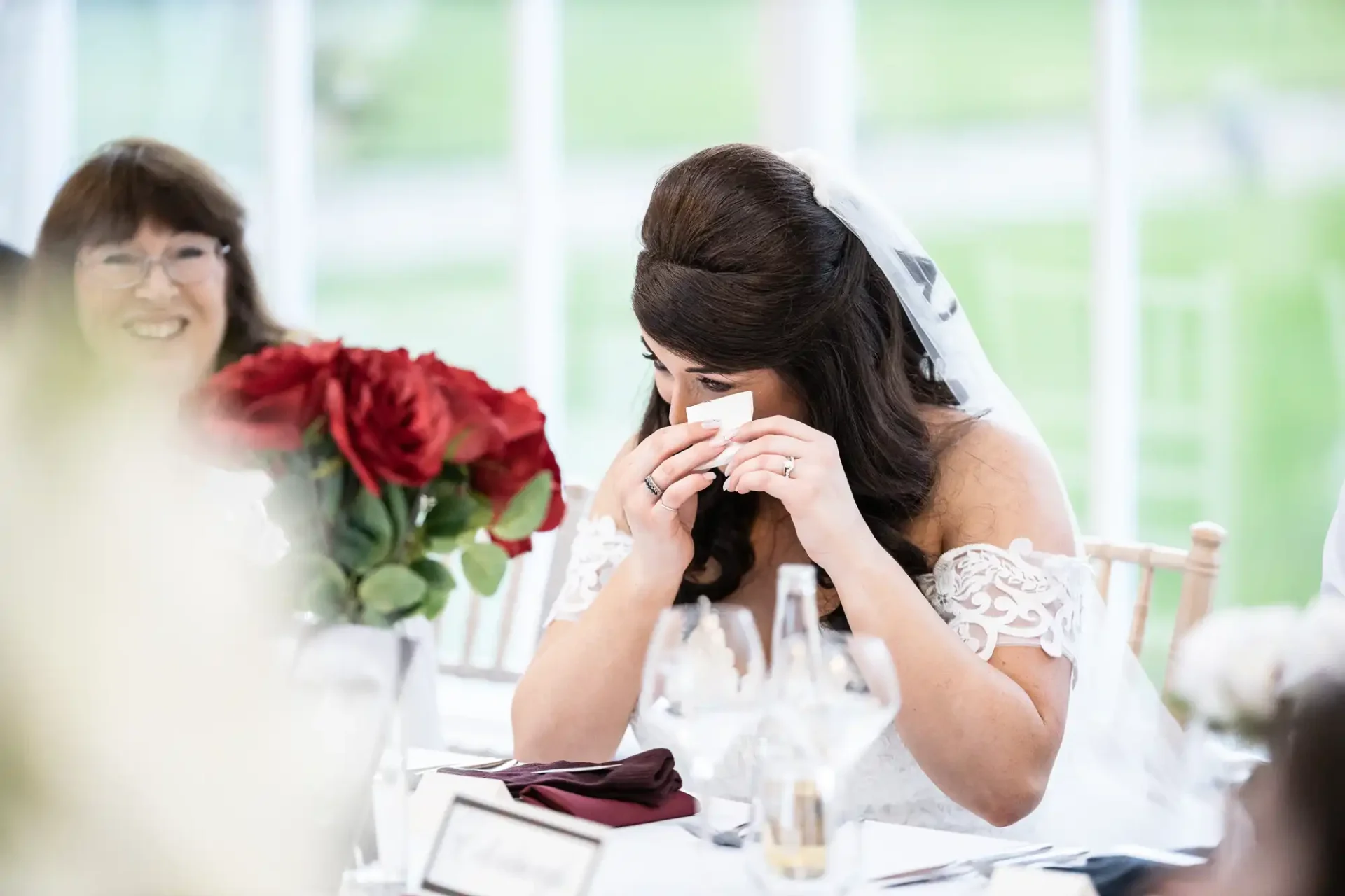 A bride in a white dress wipes tears at a wedding table, with red roses and a smiling guest nearby.
