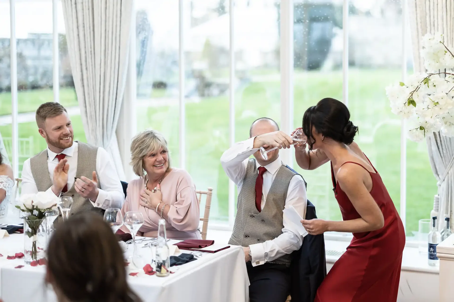 Woman in a red dress playfully covering a man's eyes at a wedding reception table while others laugh.