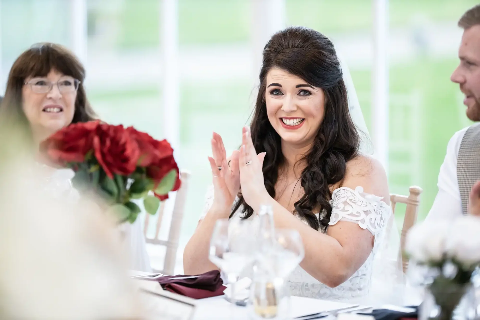 A bride smiling joyfully at a wedding table, clapping her hands, with guests and red roses nearby.