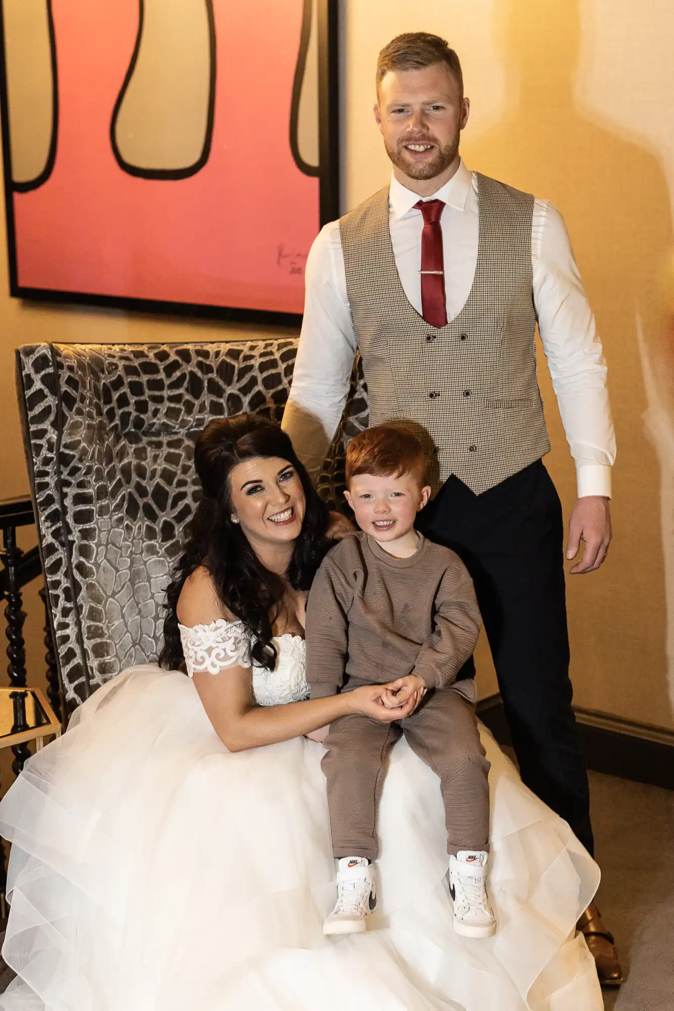 A woman in a white wedding dress sits with a young boy in brown clothes, while a man in a vest stands behind them, all smiling, in a room with artwork.