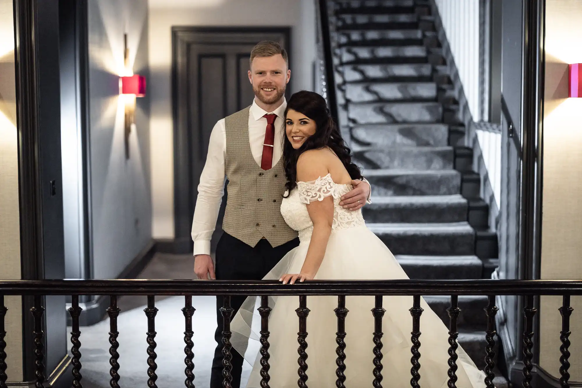 A bride in a white gown and groom in a vest and tie pose together, smiling, on a staircase landing inside a building.