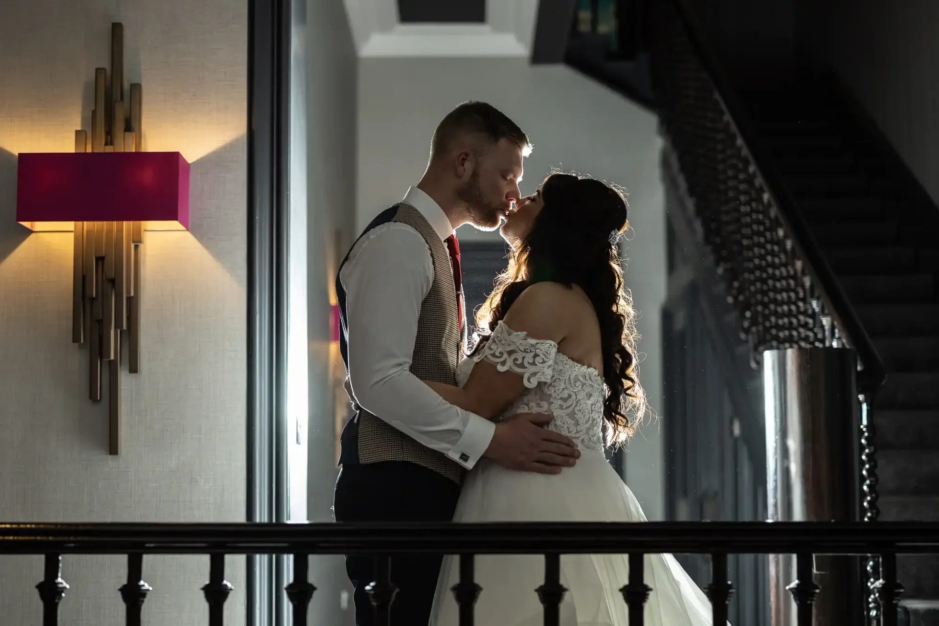 A bride and groom sharing an intimate moment on a balcony, backlit by natural light, with a staircase and lamp in the background.