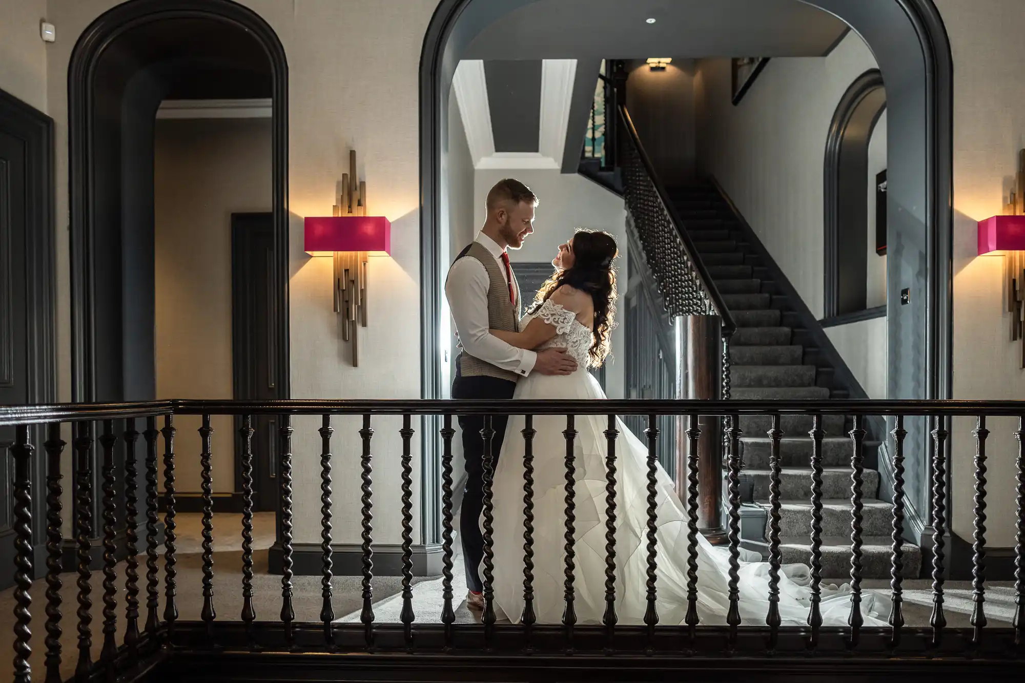 A bride and groom stand on an indoor balcony, holding each other and gazing into each other's eyes. The background features a staircase and an arched hallway with wall sconces.