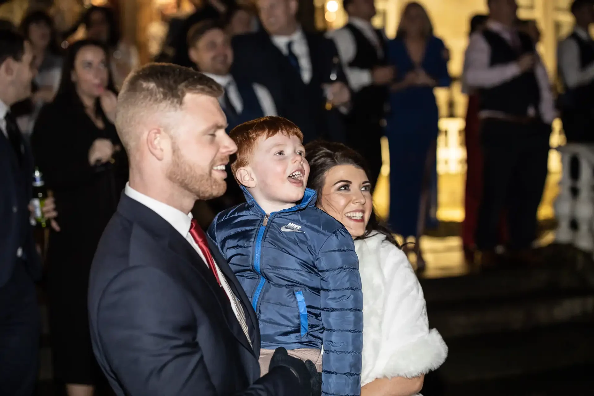 A joyful family moment at a nighttime event, featuring a man and a woman smiling while holding a small boy in a blue jacket. other attendees are visible in the background.