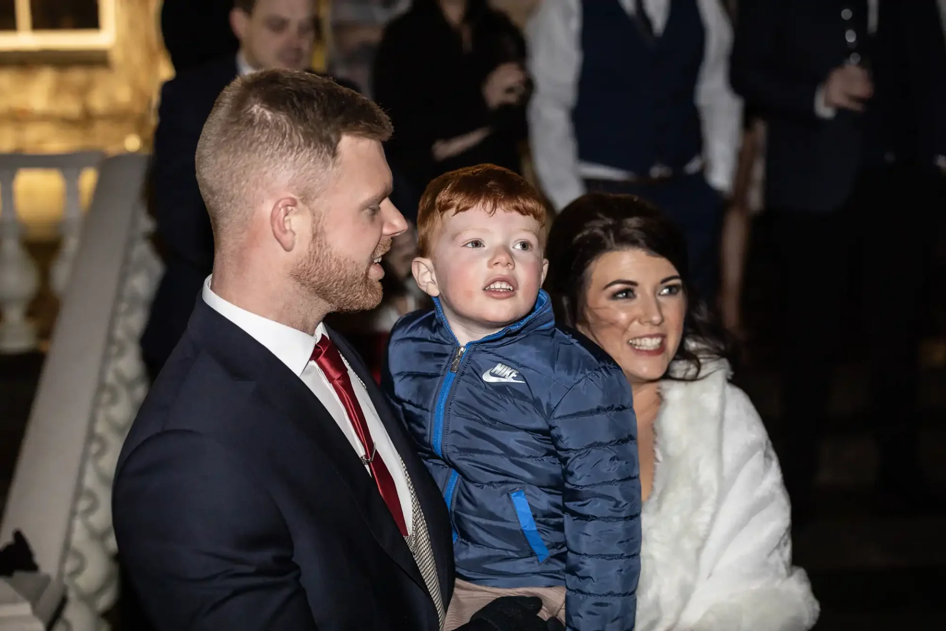 A family at a night event, with a man in a suit and a woman in a white fur coat holding a young boy in a blue jacket. they are smiling and looking up.
