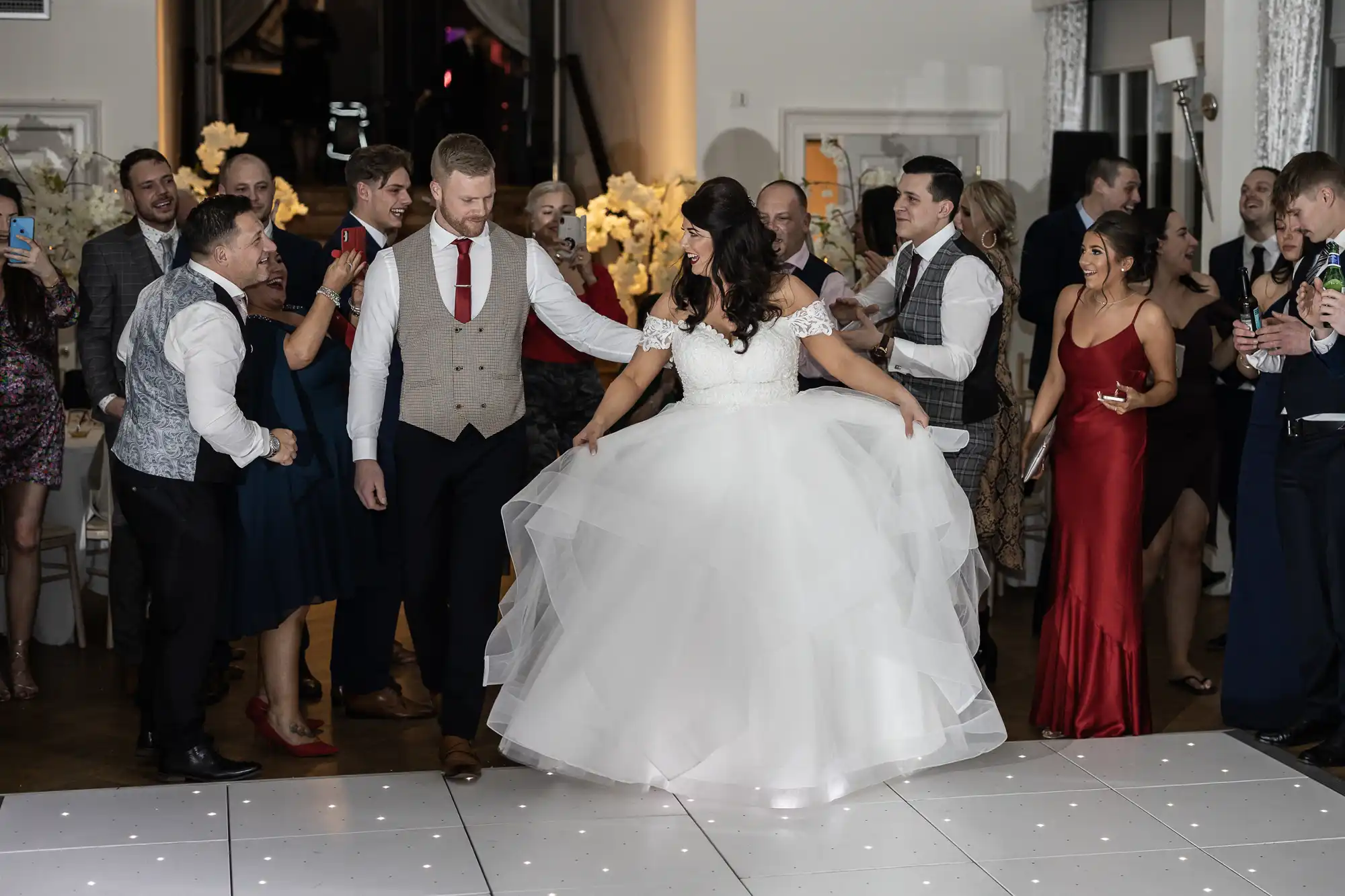 A couple dances at their wedding reception, surrounded by guests in formal attire who are clapping and watching them on a white-lit dance floor.
