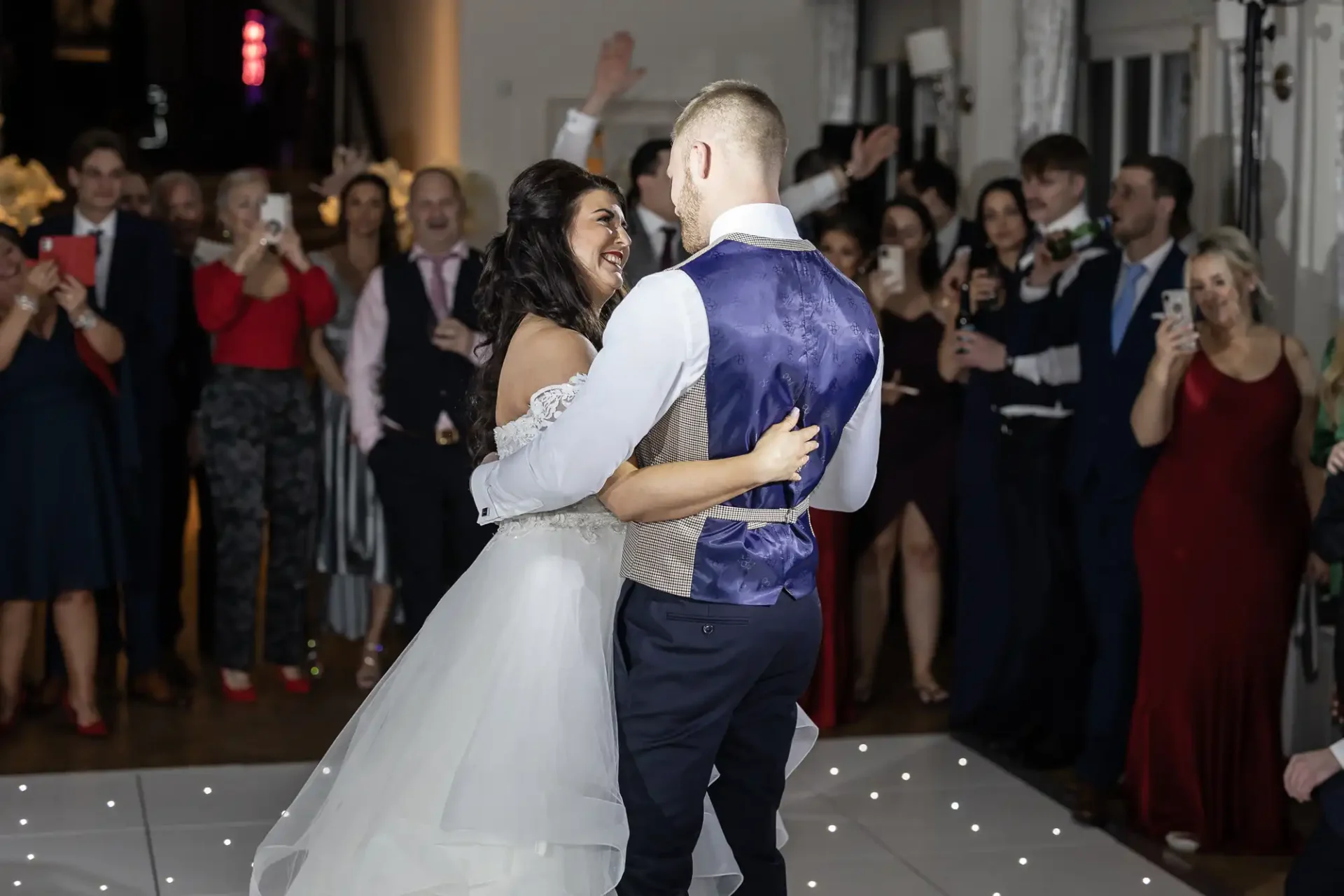 A newlywed couple dances joyfully at their wedding reception surrounded by applauding guests.