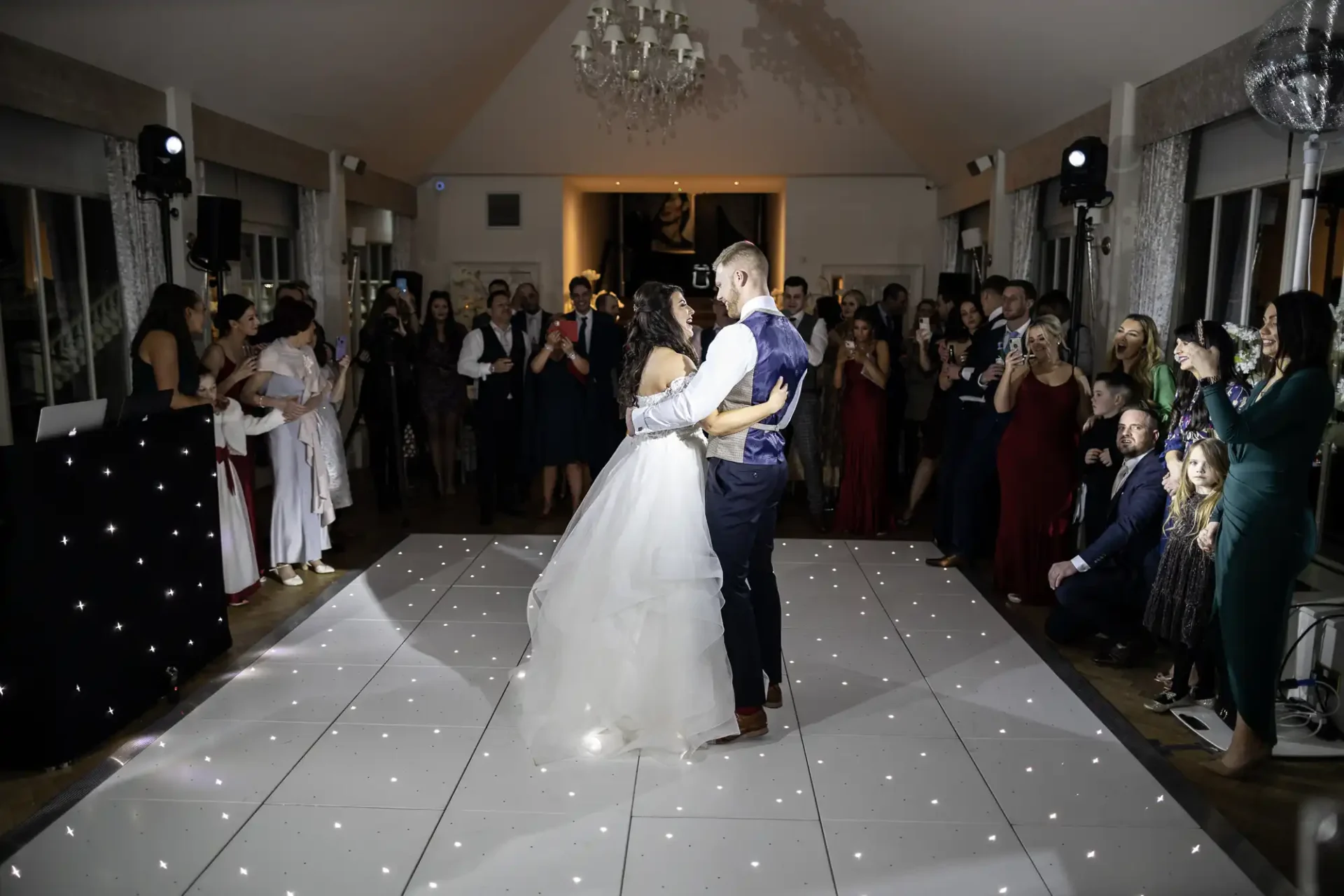 A bride and groom share a dance on a lit dance floor surrounded by guests at a wedding reception.