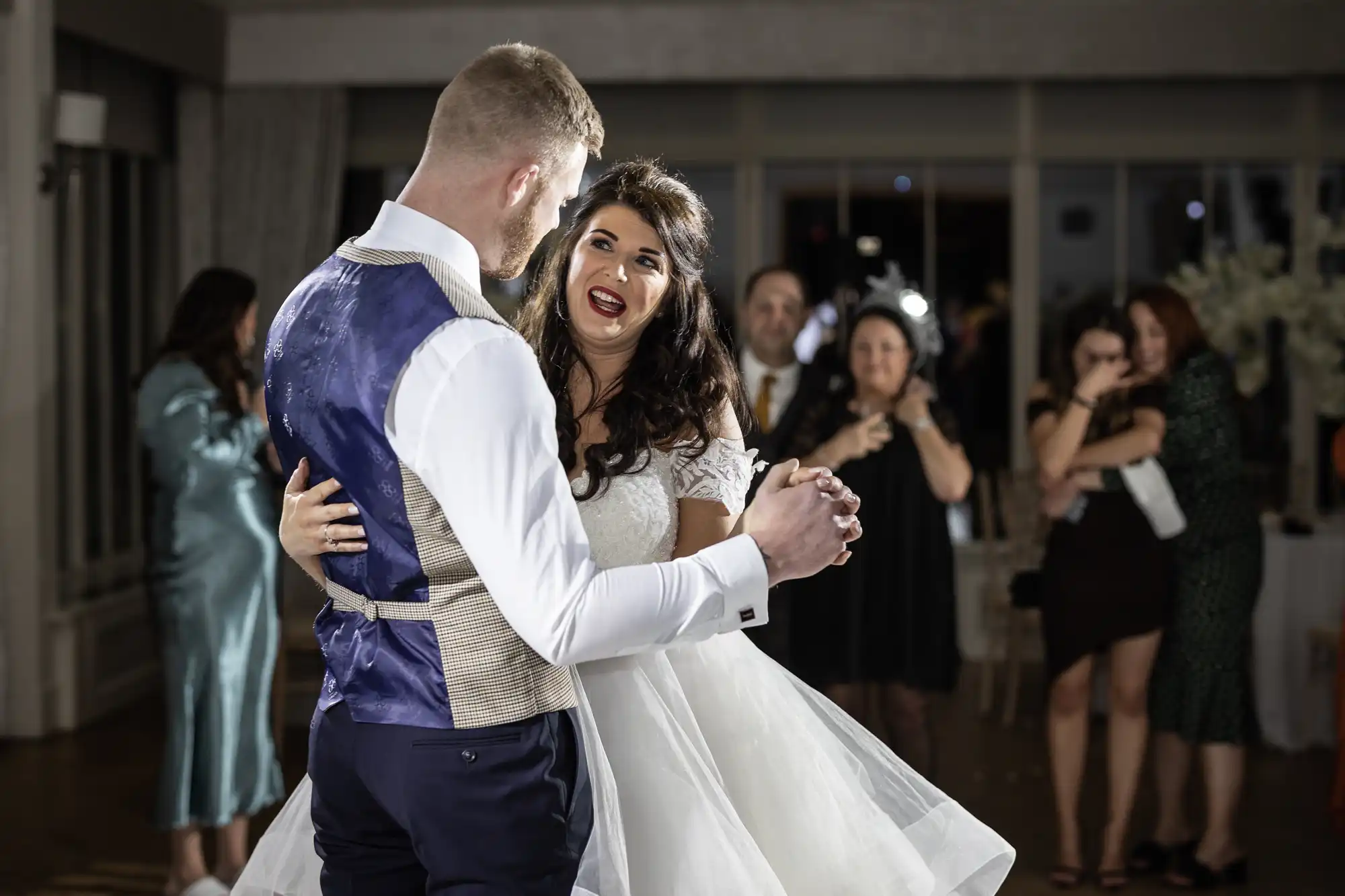 A bride and groom share a dance at their wedding reception, with guests watching and taking photos in the background.