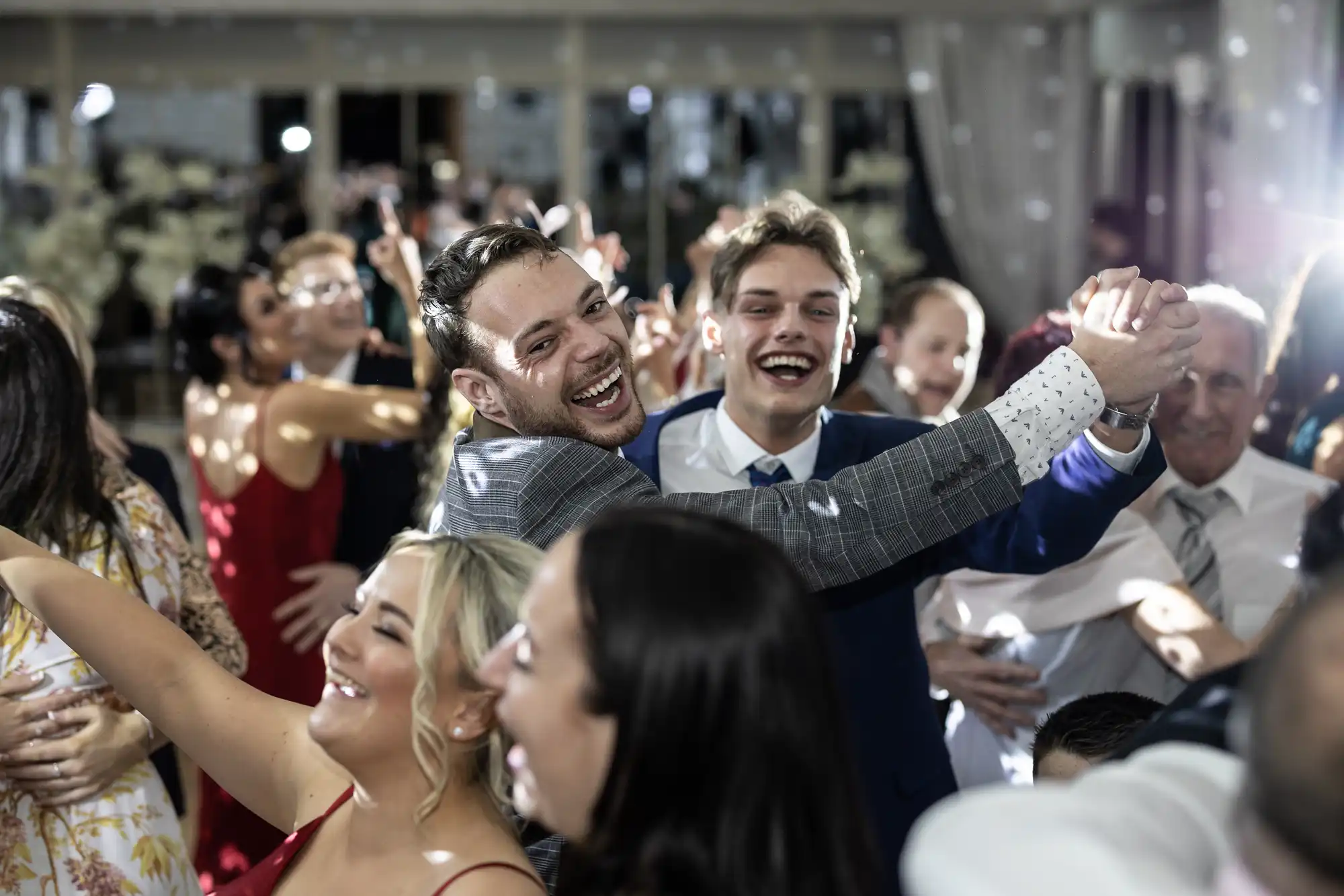 A group of people, including two men in suits at the center, smile and dance together in a lively indoor setting.