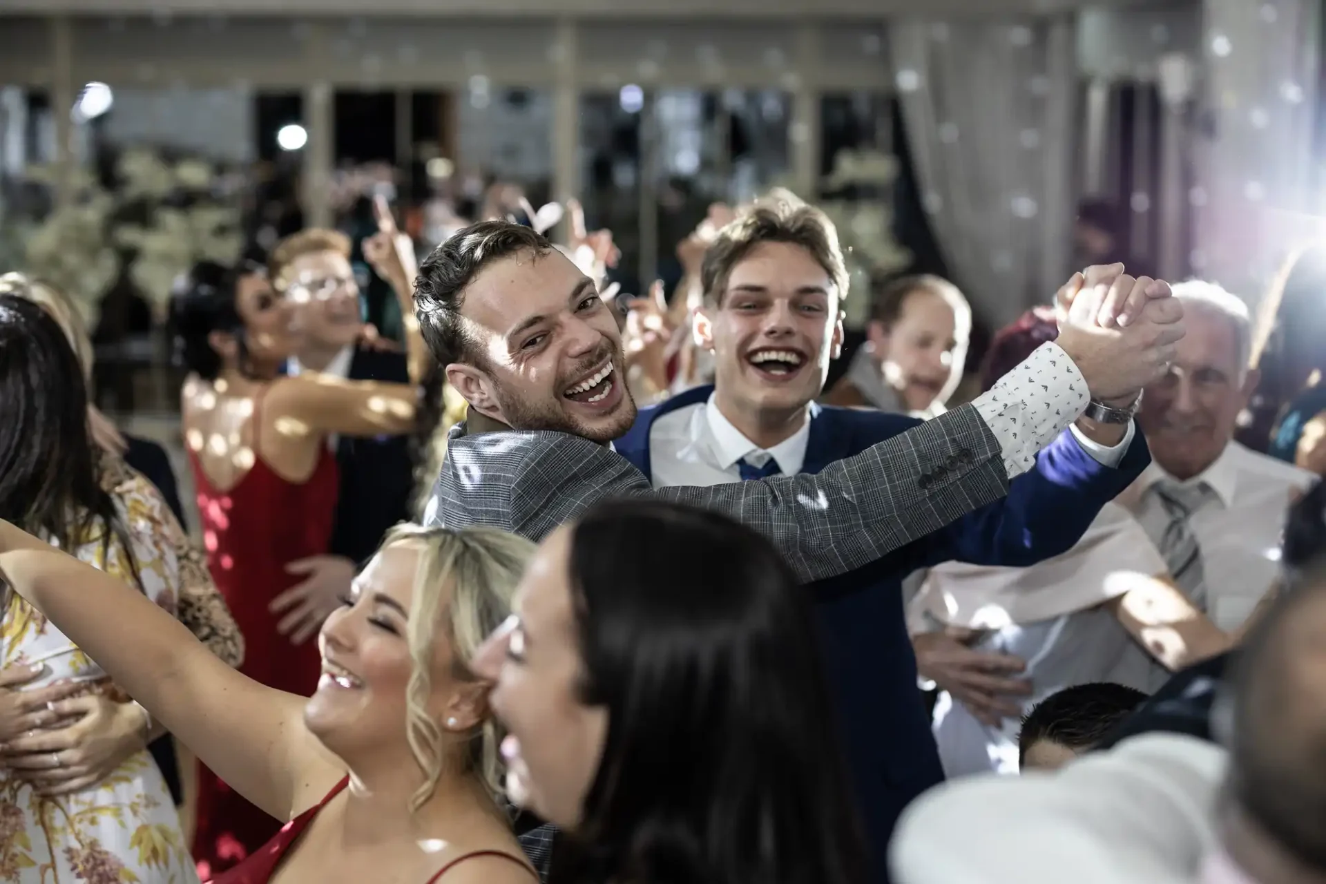 Two joyful young men dancing and taking a selfie at a festive gathering with other guests smiling around them.