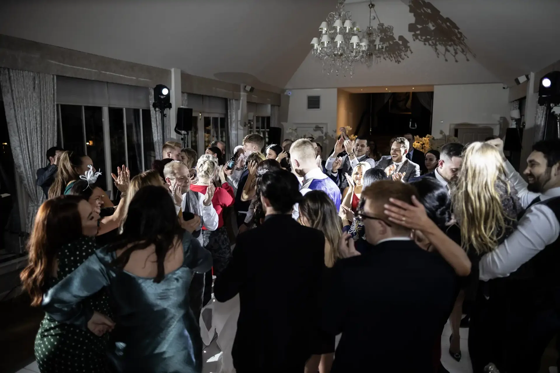 Guests dancing enthusiastically at a lively indoor evening reception, with a chandelier overhead and dj equipment visible.