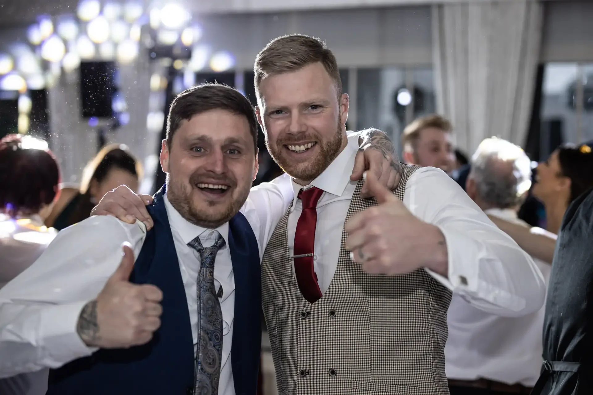 Two joyful men in formal attire giving thumbs up at a festive event with blurred guests and lights in the background.