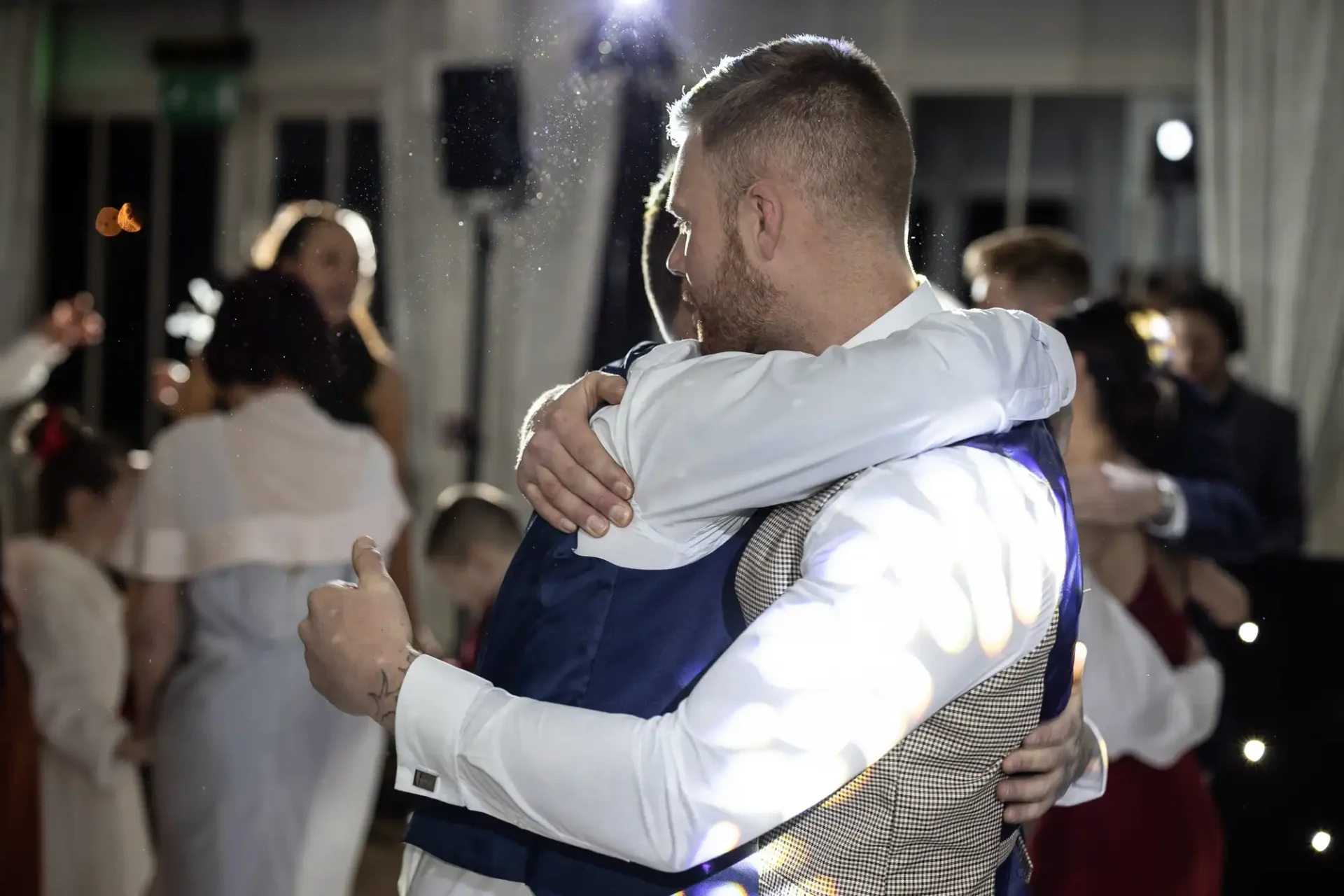 Two men embracing at a wedding reception, one giving a thumbs-up, with guests in the background and a flash of light capturing floating particles in the air.