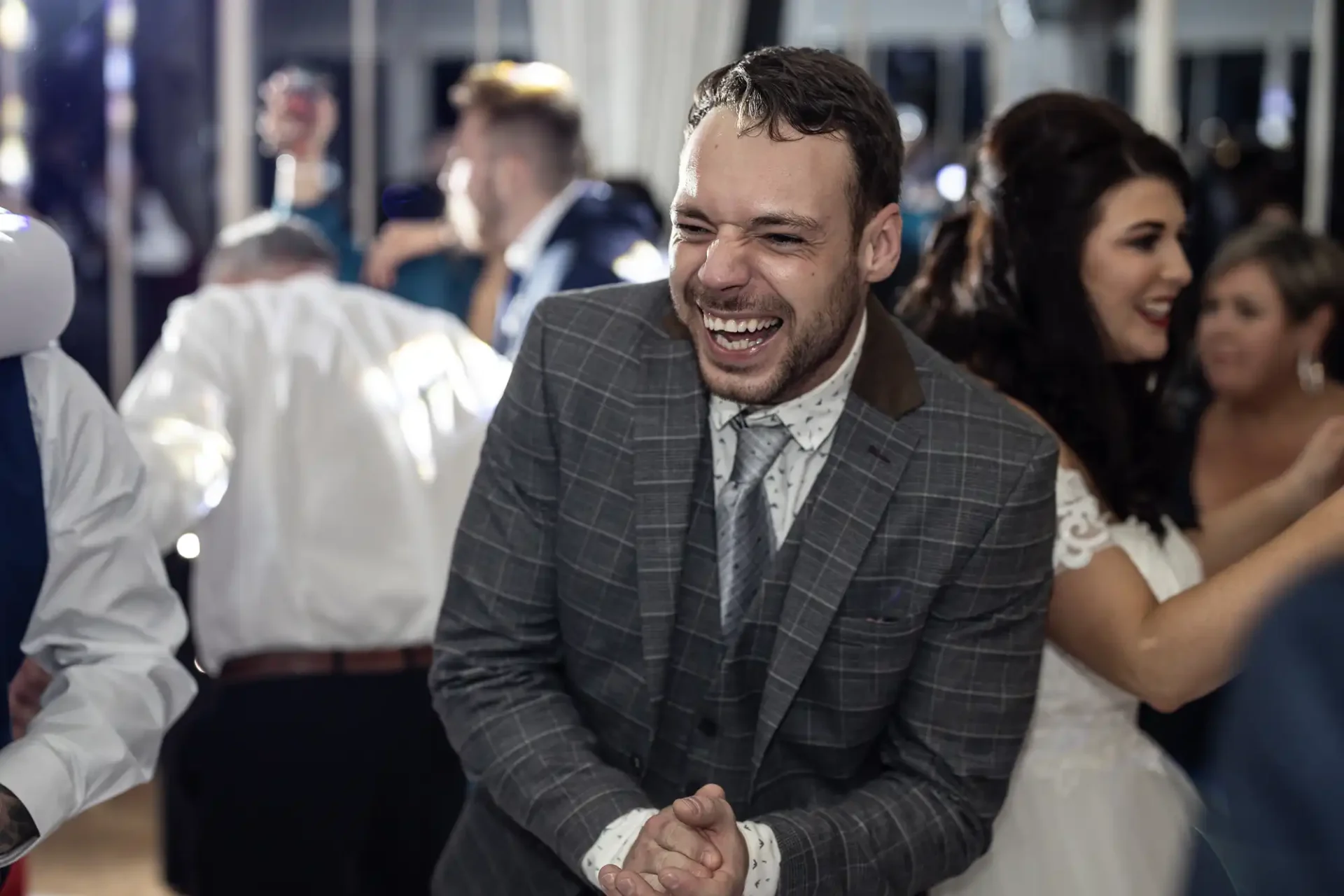 Laughing man in a gray suit claps hands at a wedding reception, with other guests and a bride in the background.