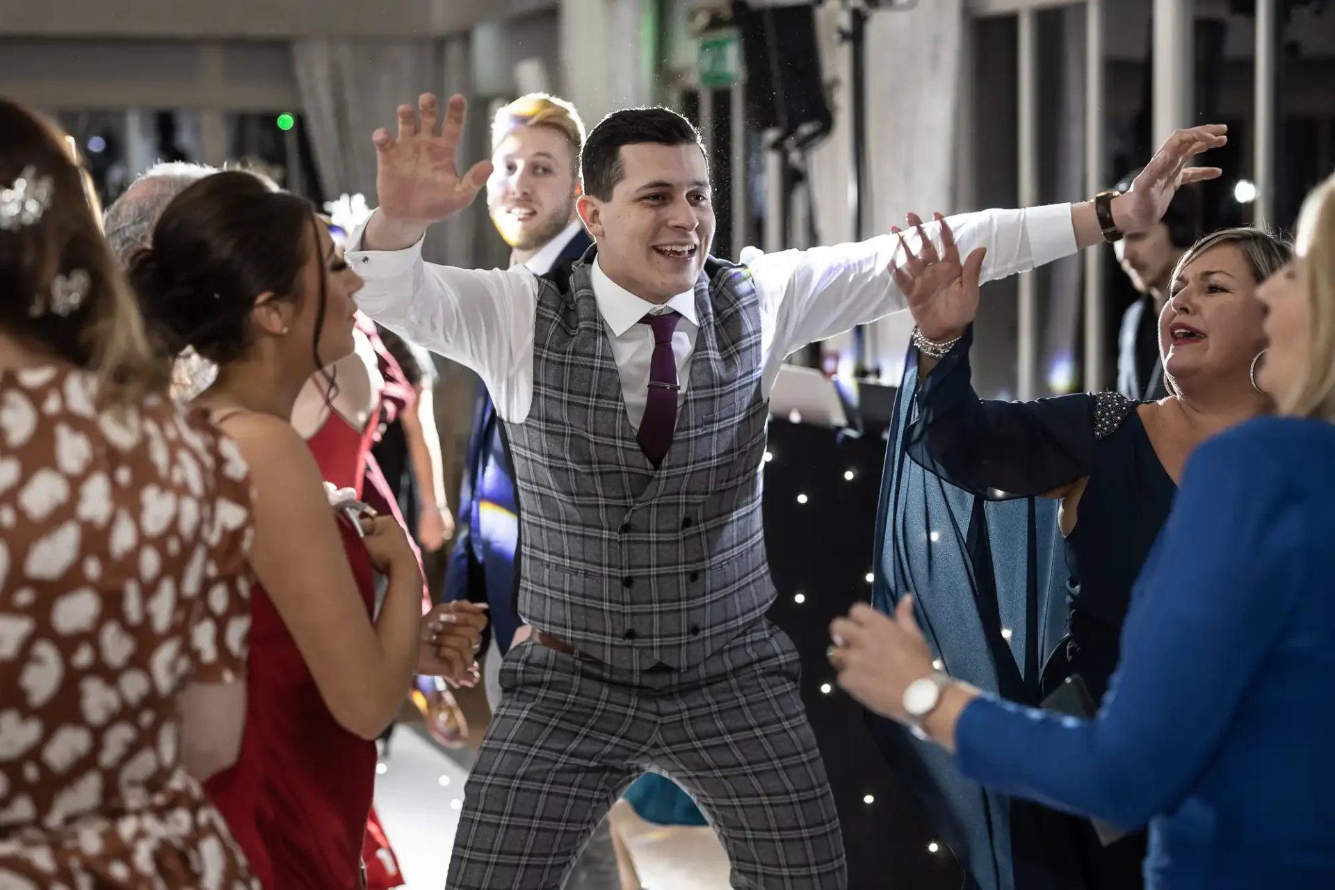 A man in a plaid suit dancing joyfully with raised arms among a group of elegantly dressed people at a lively indoor party.