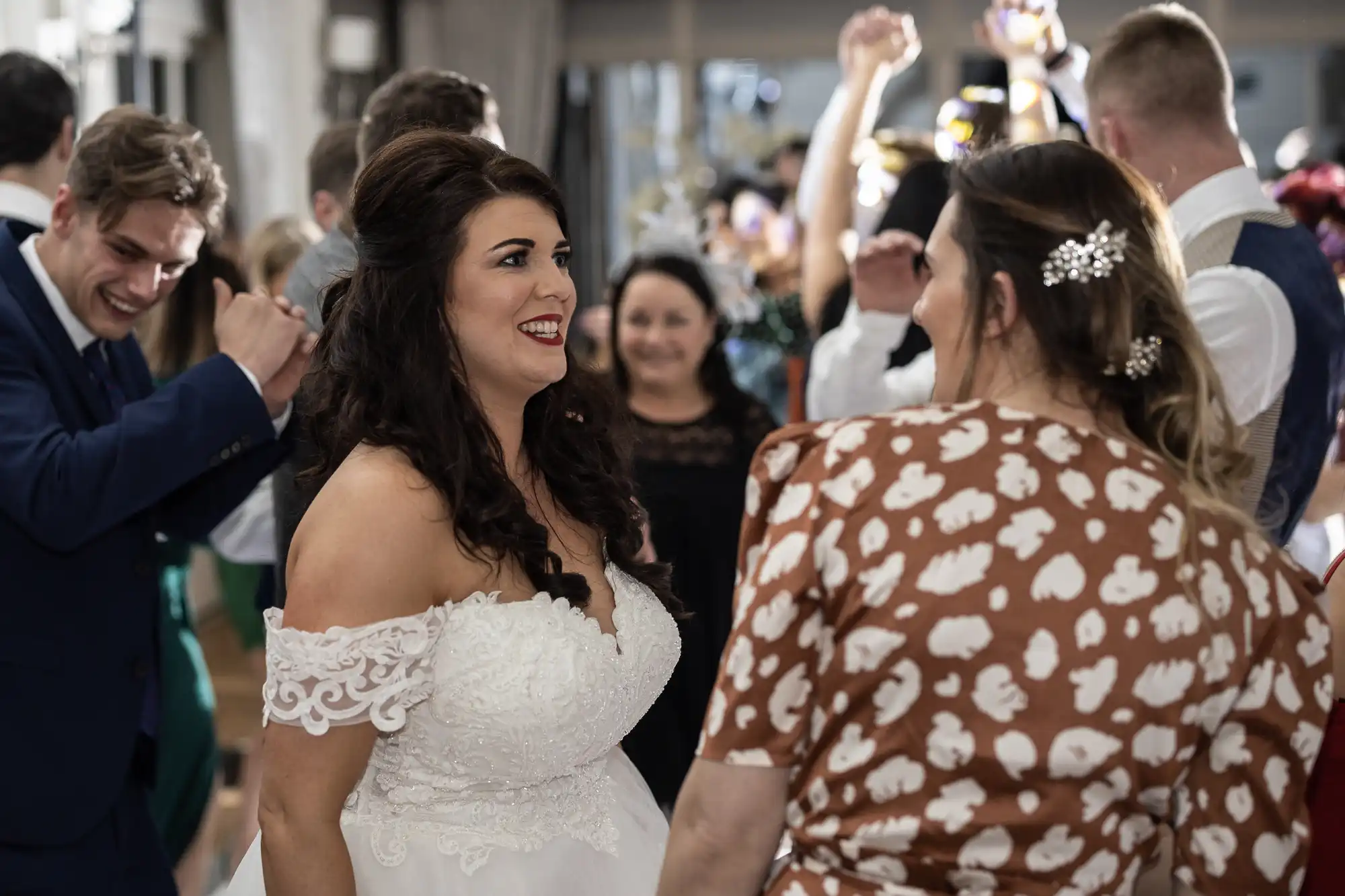 A bride in a white dress is smiling and talking to a guest in a brown patterned dress at a wedding reception. People are dancing in the background.